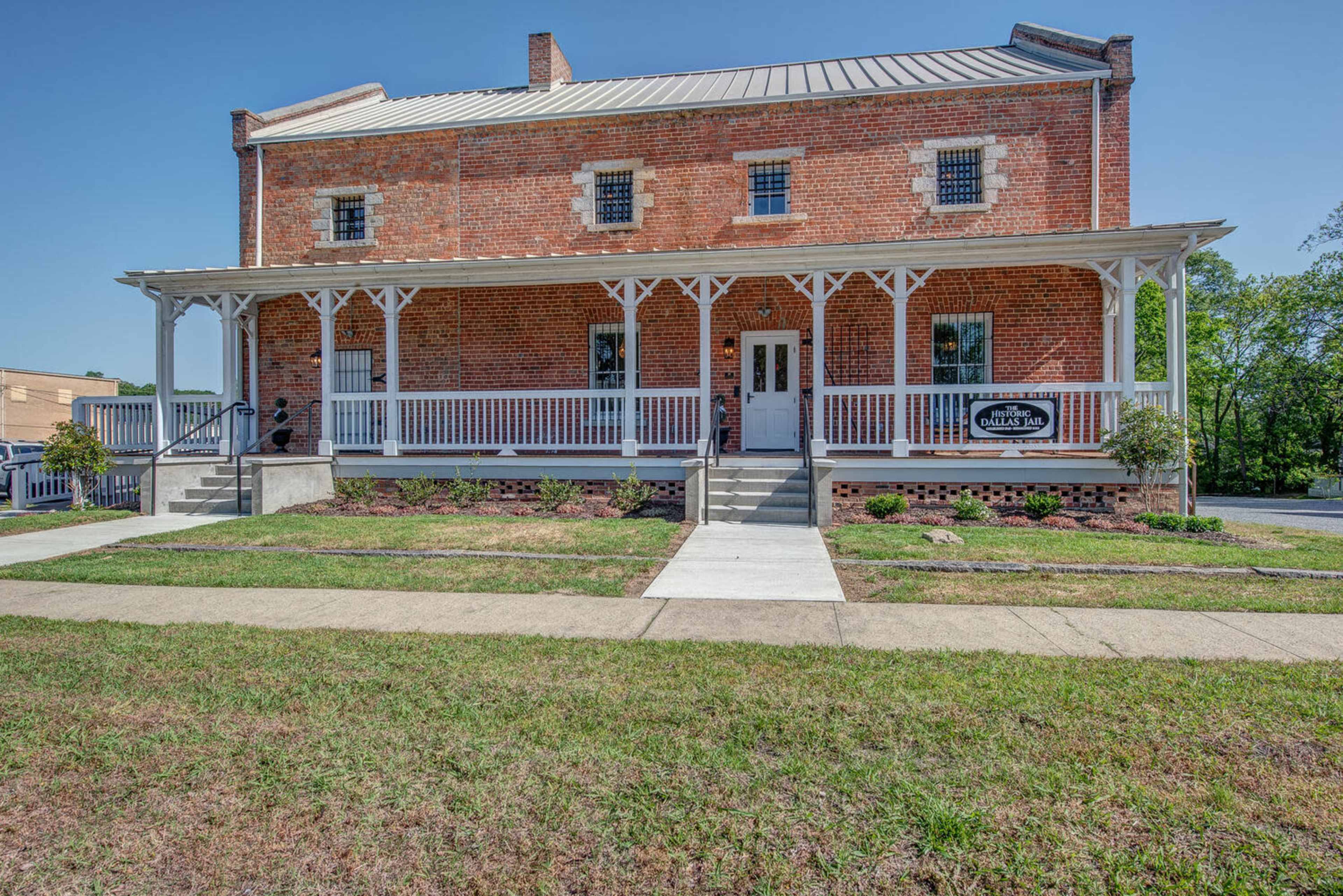 A two-story brick building with a front porch and white railings, located on a grassy area with a sidewalk leading to the entrance.
