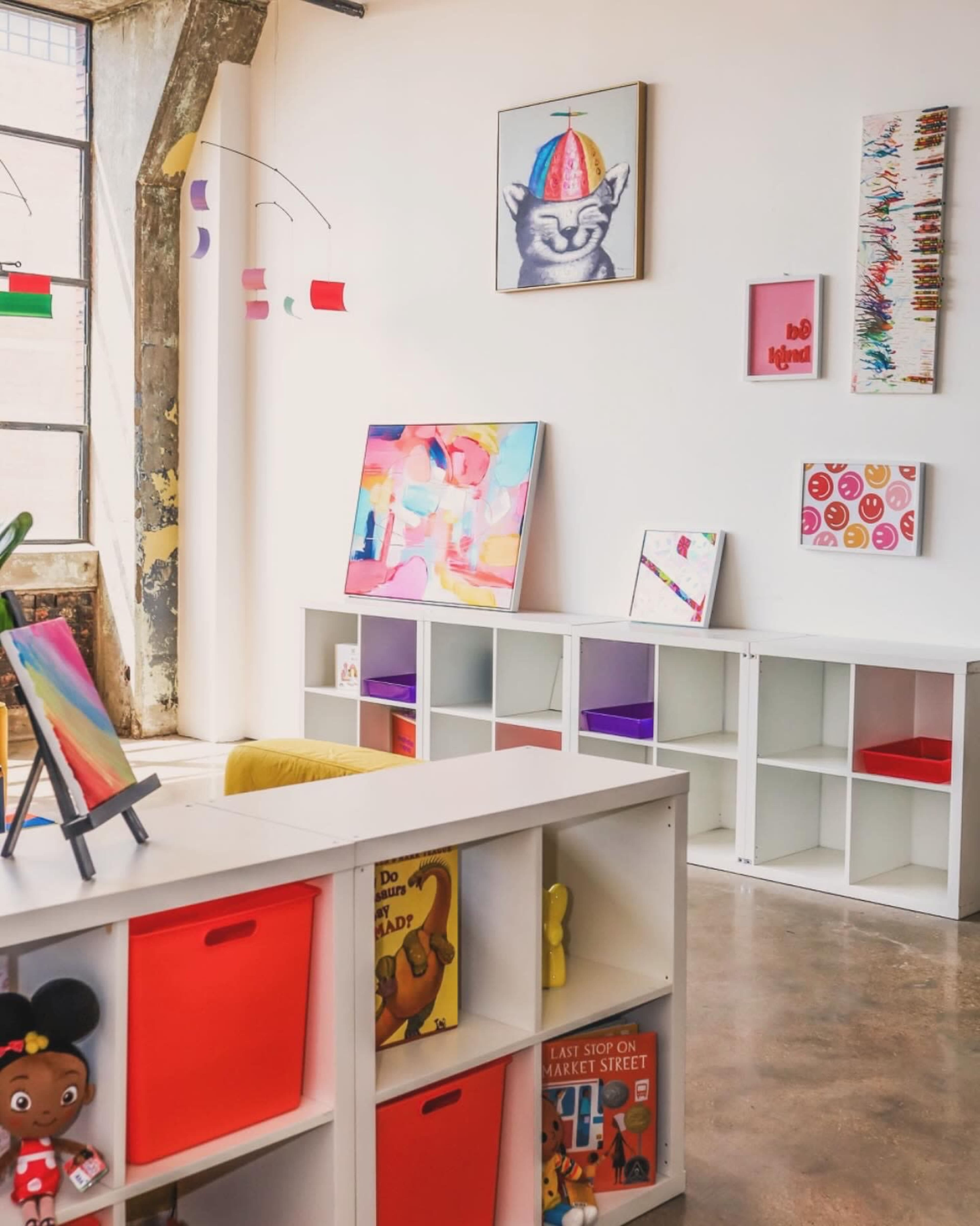A brightly lit room with white walls adorned with colorful artwork and a white shelving unit containing red and orange storage bins alongside books and toys.