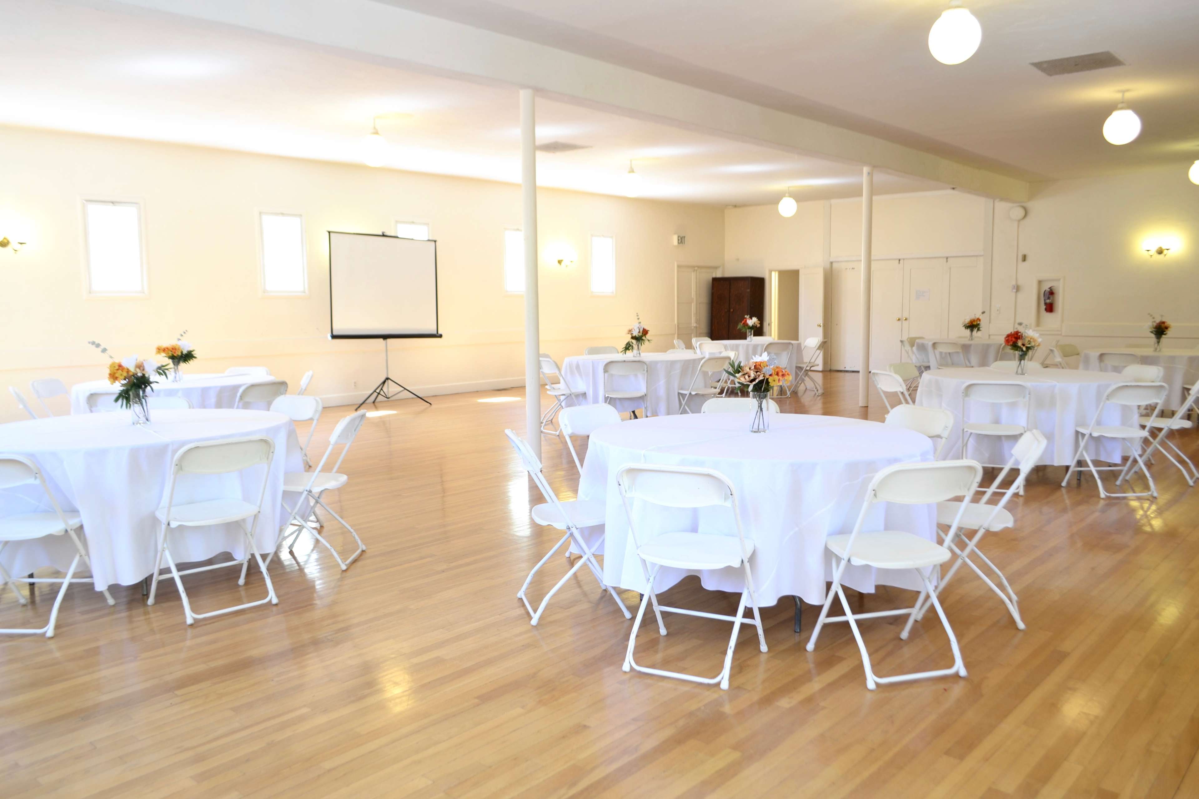 A spacious, well-lit hall arranged with several round tables covered in white tablecloths, each adorned with small flower centerpieces.