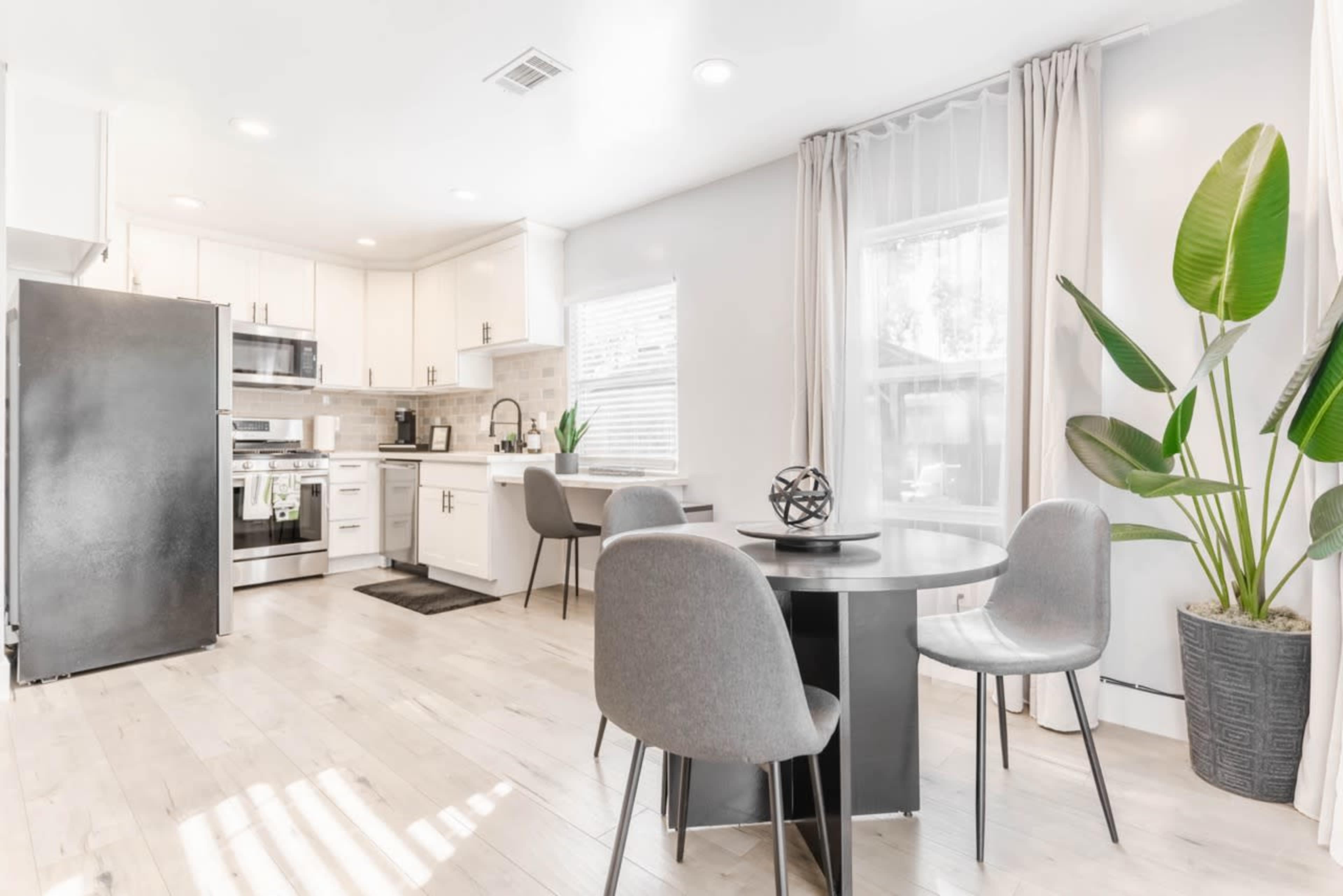 A modern kitchen and dining area features white cabinetry, stainless steel appliances, a round table with gray chairs, and a large potted plant by the window.