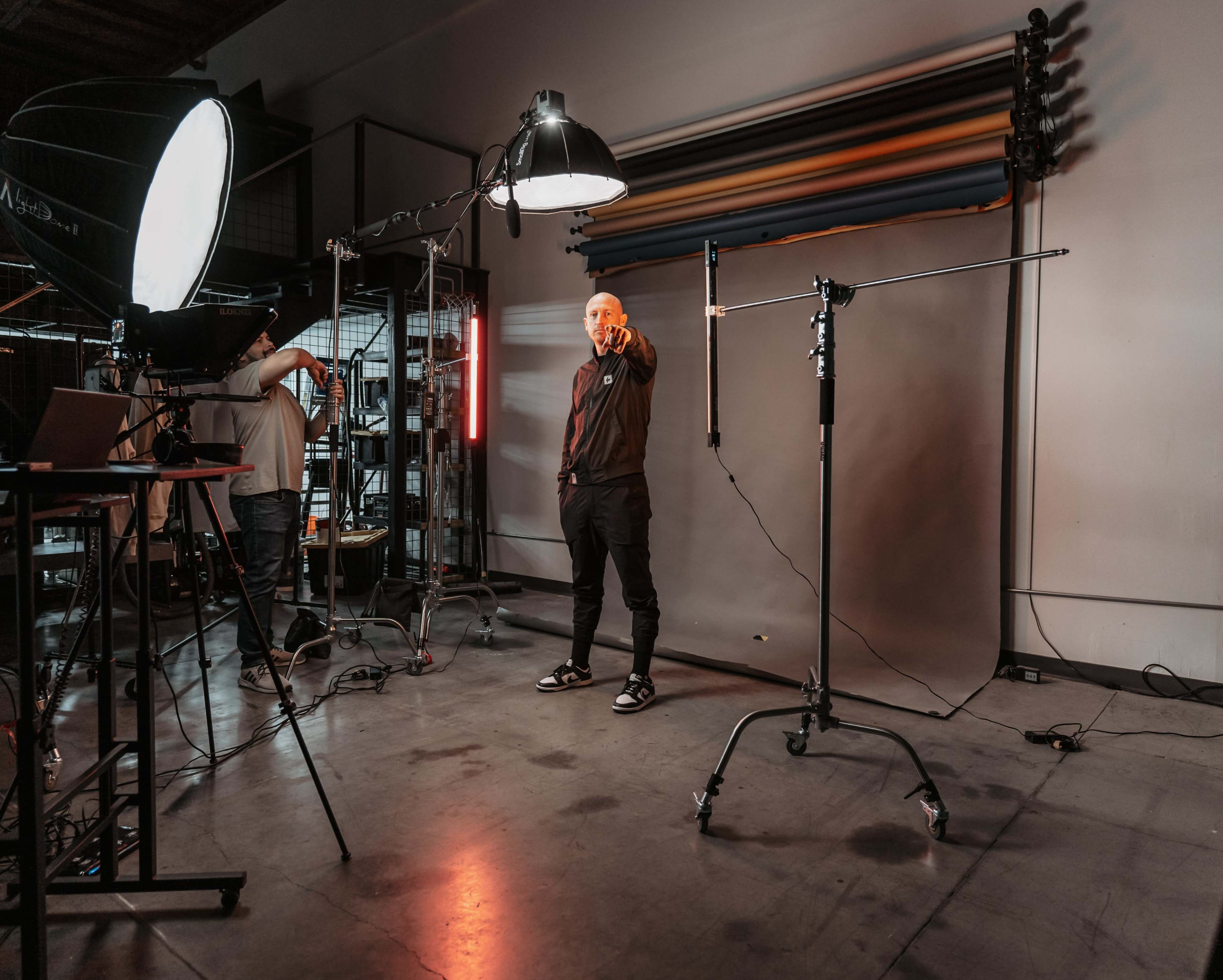 A person stands in a photography studio, pointing towards the camera while surrounded by lighting equipment and a backdrop.