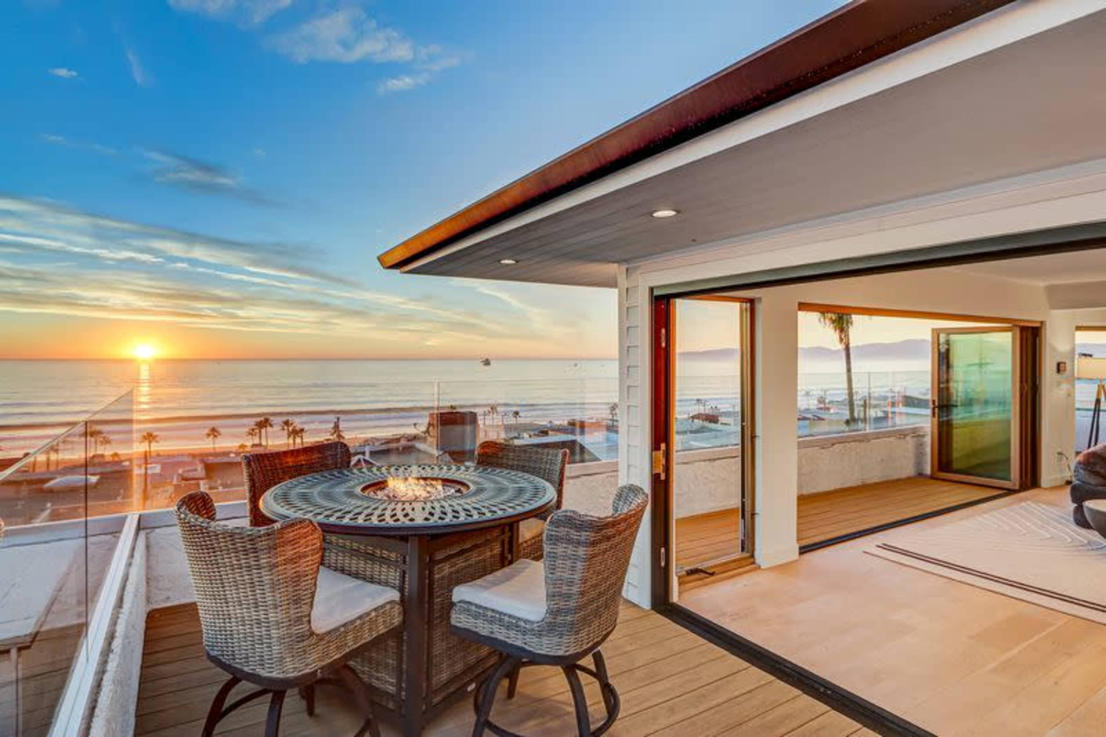 The image shows a modern terrace with a dining table and chairs overlooking a sunset view of the ocean and coastline.