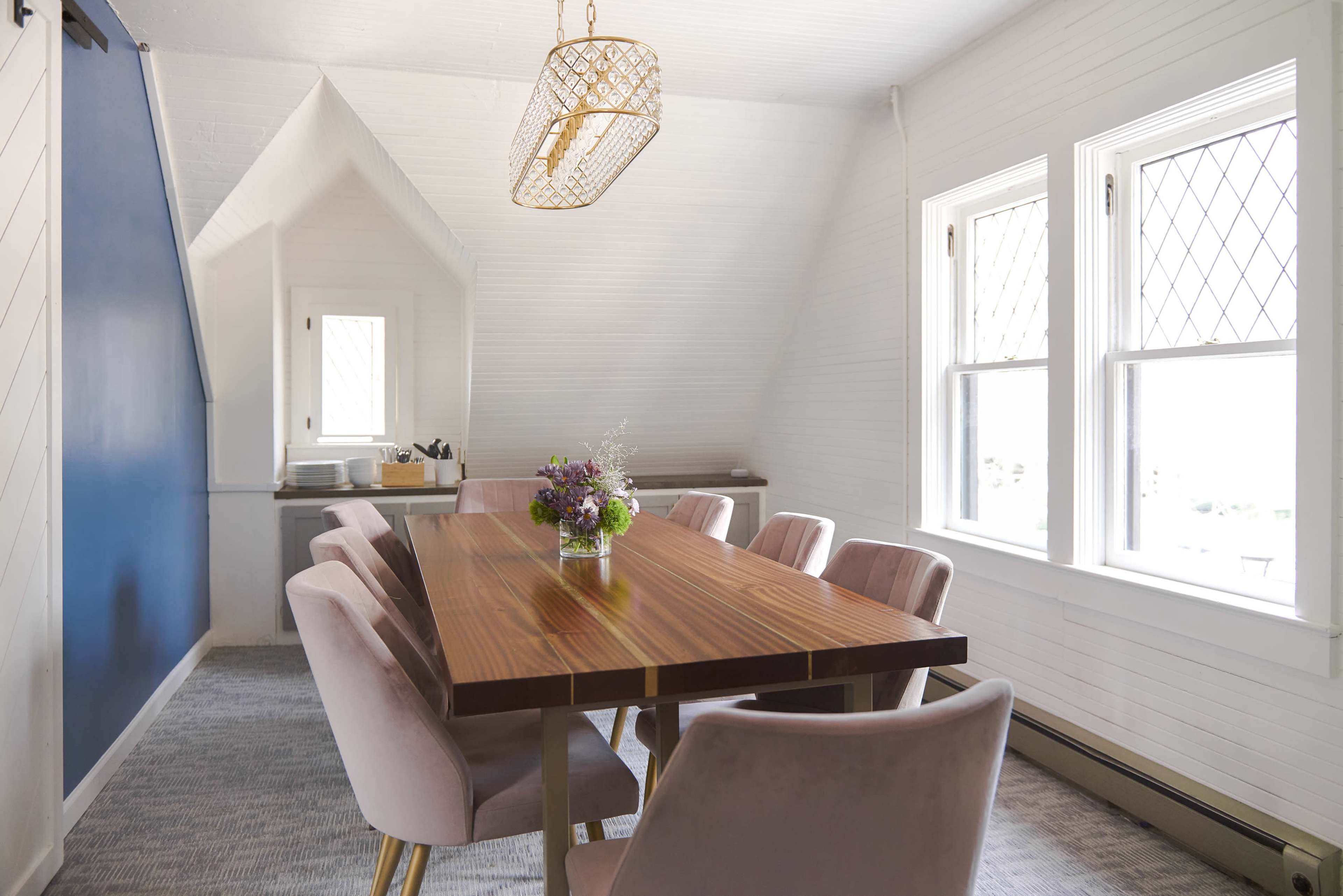 A modern dining room features a long wooden table surrounded by pink upholstered chairs, illuminated by a chandelier, with large windows letting in natural light.