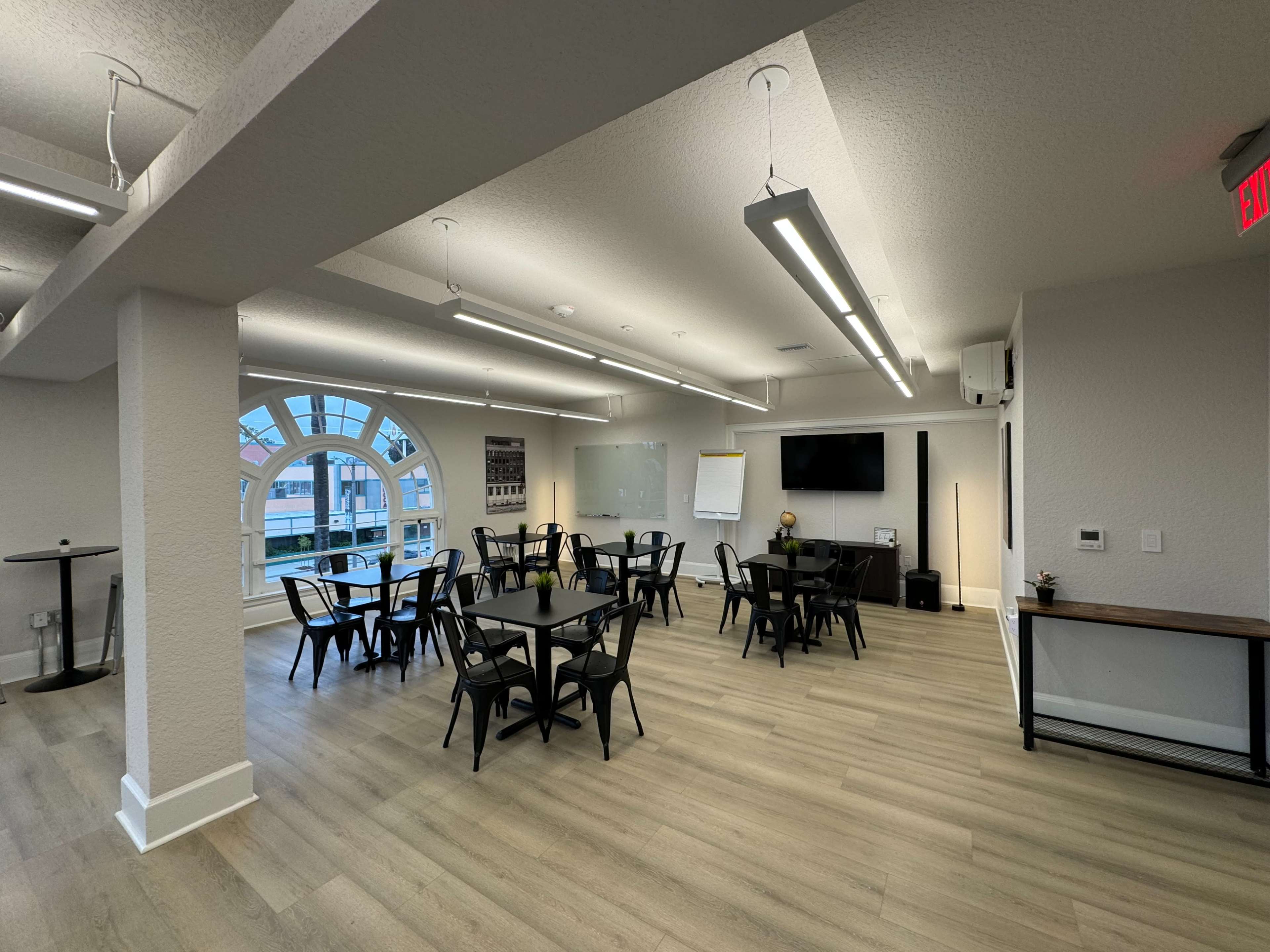 A modern meeting room with black chairs and tables arranged neatly, large windows allowing natural light to enter, and a wall-mounted screen.