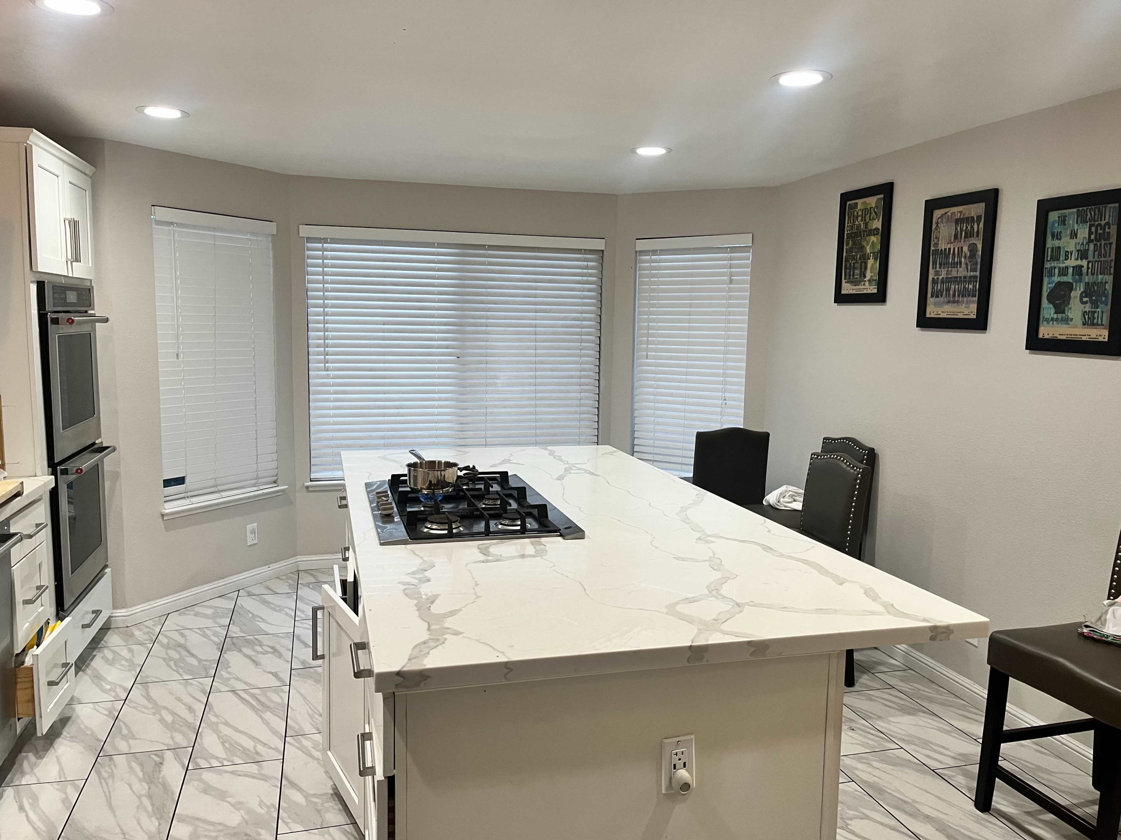 A modern kitchen with a large island featuring a gas stovetop, surrounded by windows with blinds and framed artwork on the walls.
