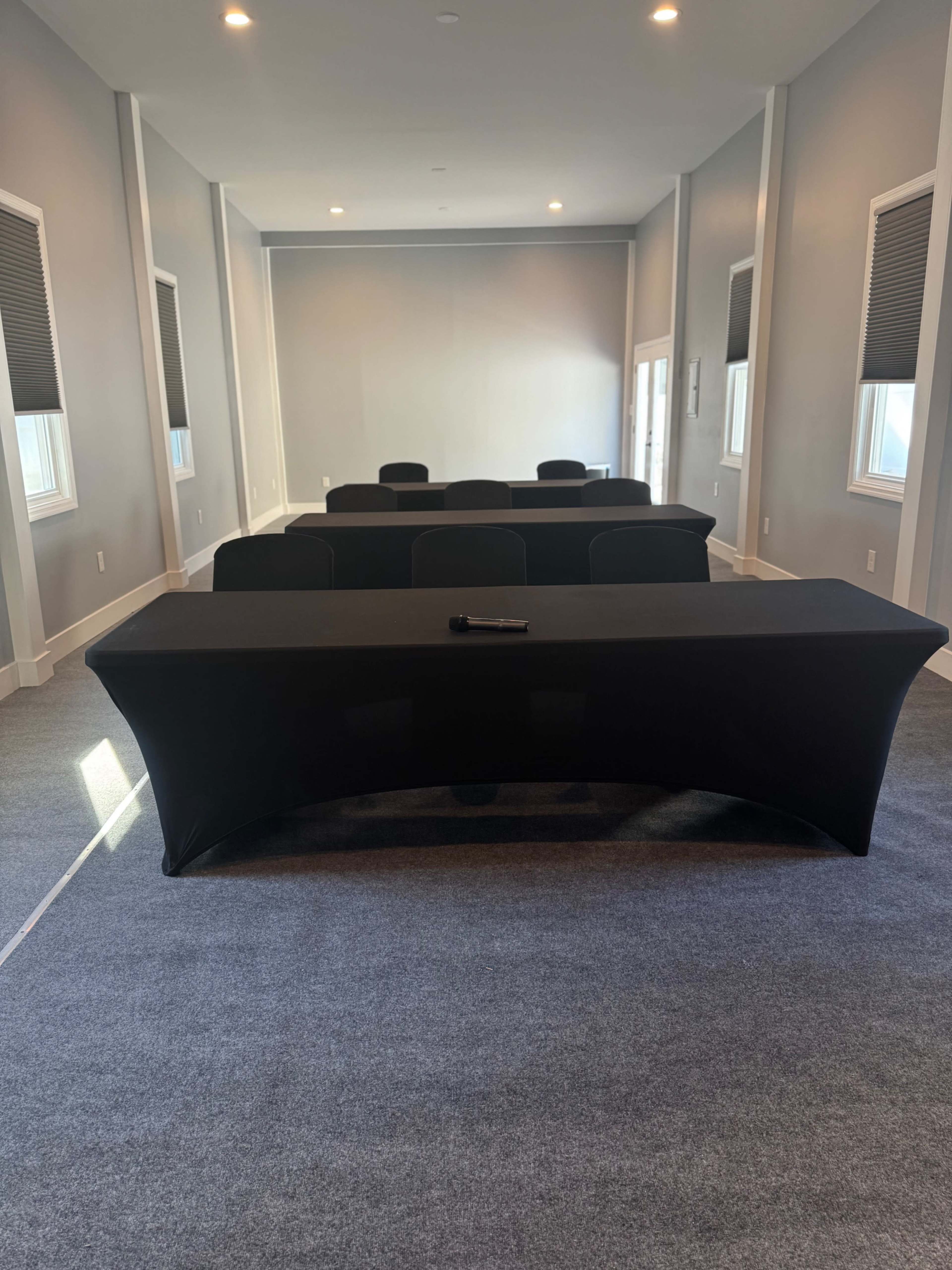 The image shows a conference room with a long table covered in a black tablecloth, surrounded by several chairs, and illuminated by natural light coming through windows.