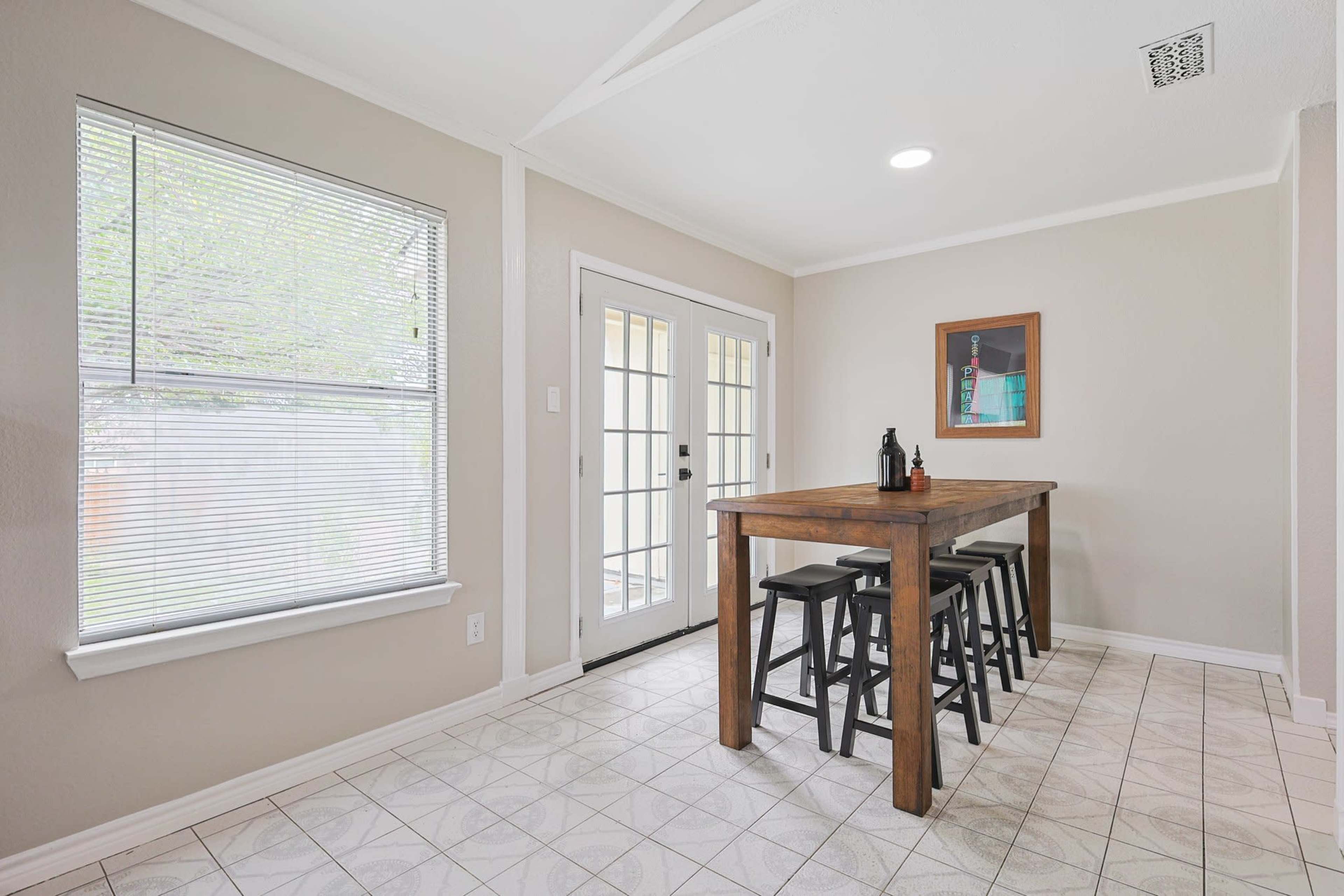 The image shows a dining area with a wooden table and four black stools, alongside a door leading to an outdoor space and a window allowing natural light to enter.