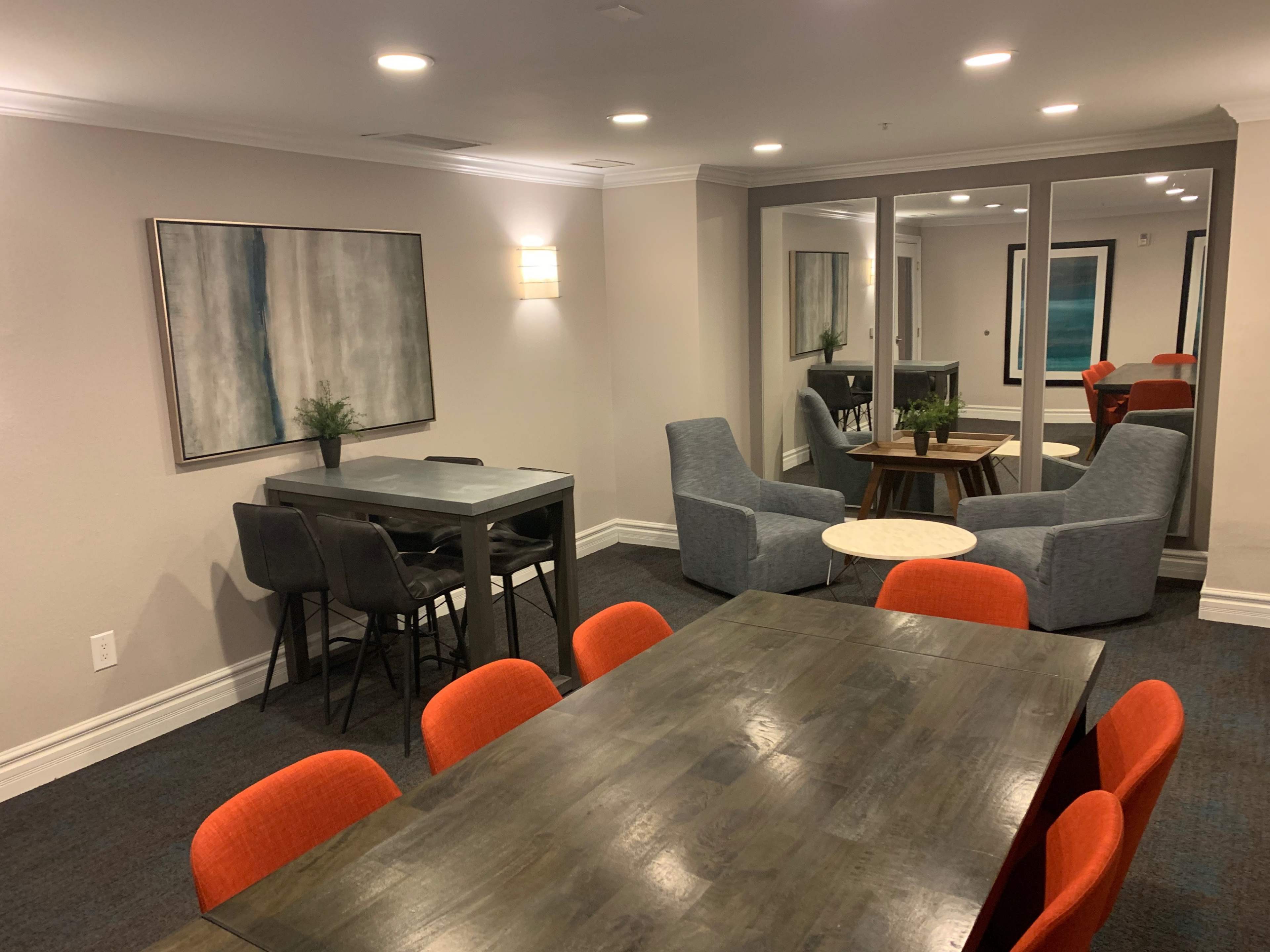 The image shows a modern lounge area featuring two seating arrangements, a large wooden table surrounded by orange chairs, and two gray armchairs near a small round table.