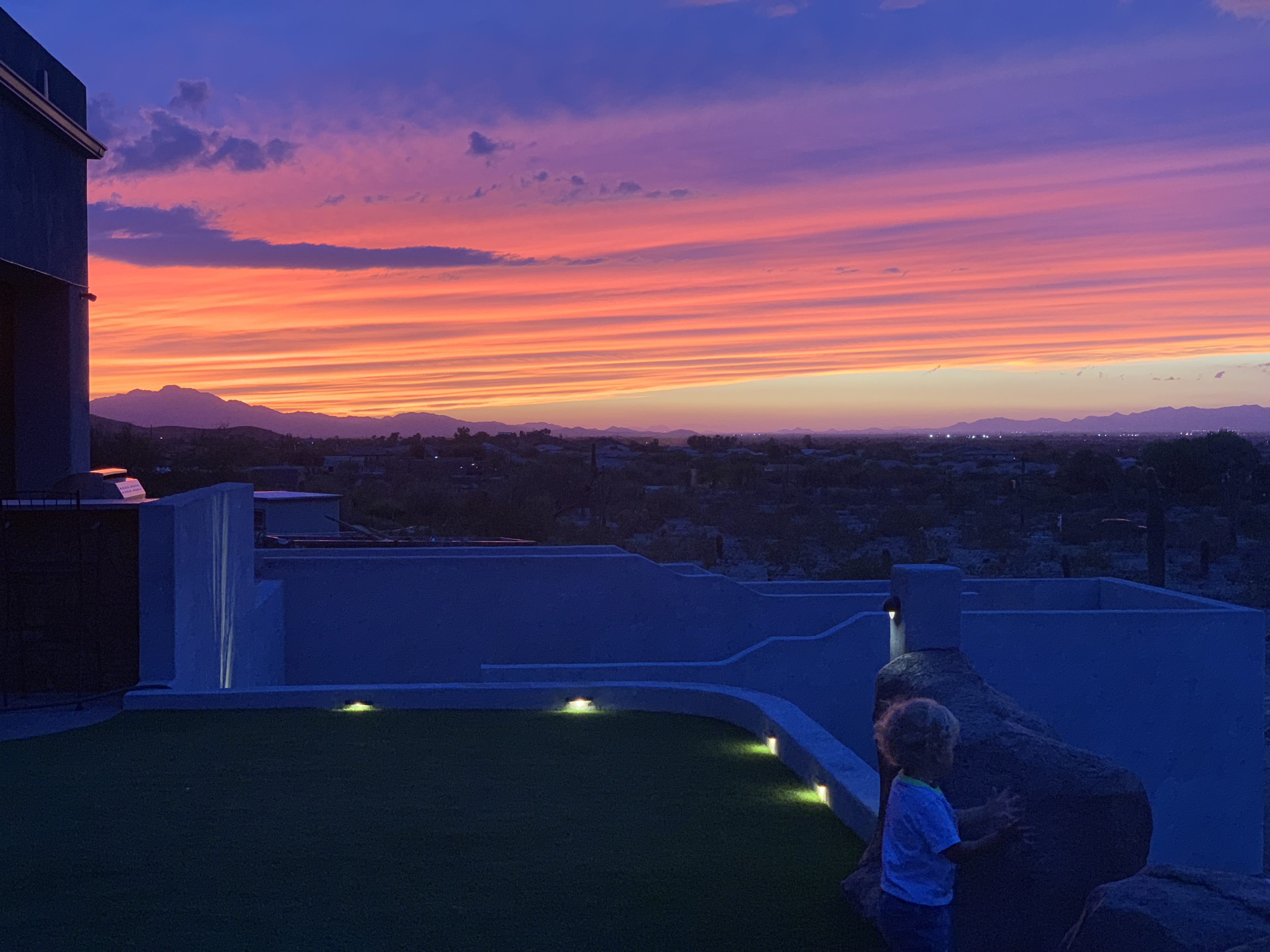 A child stands on a grassy area of a modern property, observing a colorful sunset with layered clouds over distant mountains.
