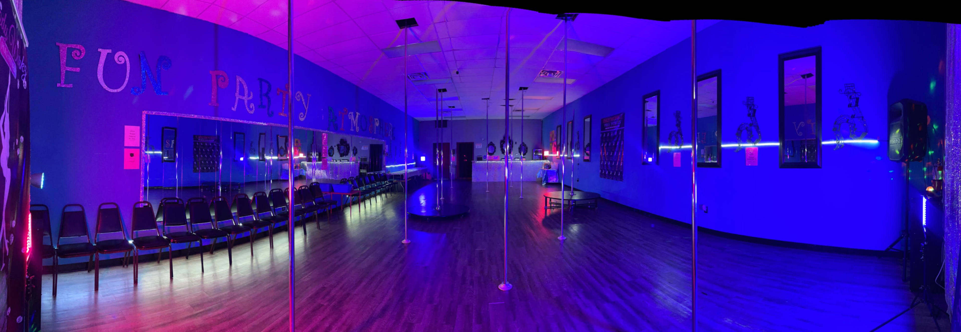 The image shows a brightly lit dance studio with mirrored walls, a central pole, and rows of black chairs along the sides.
