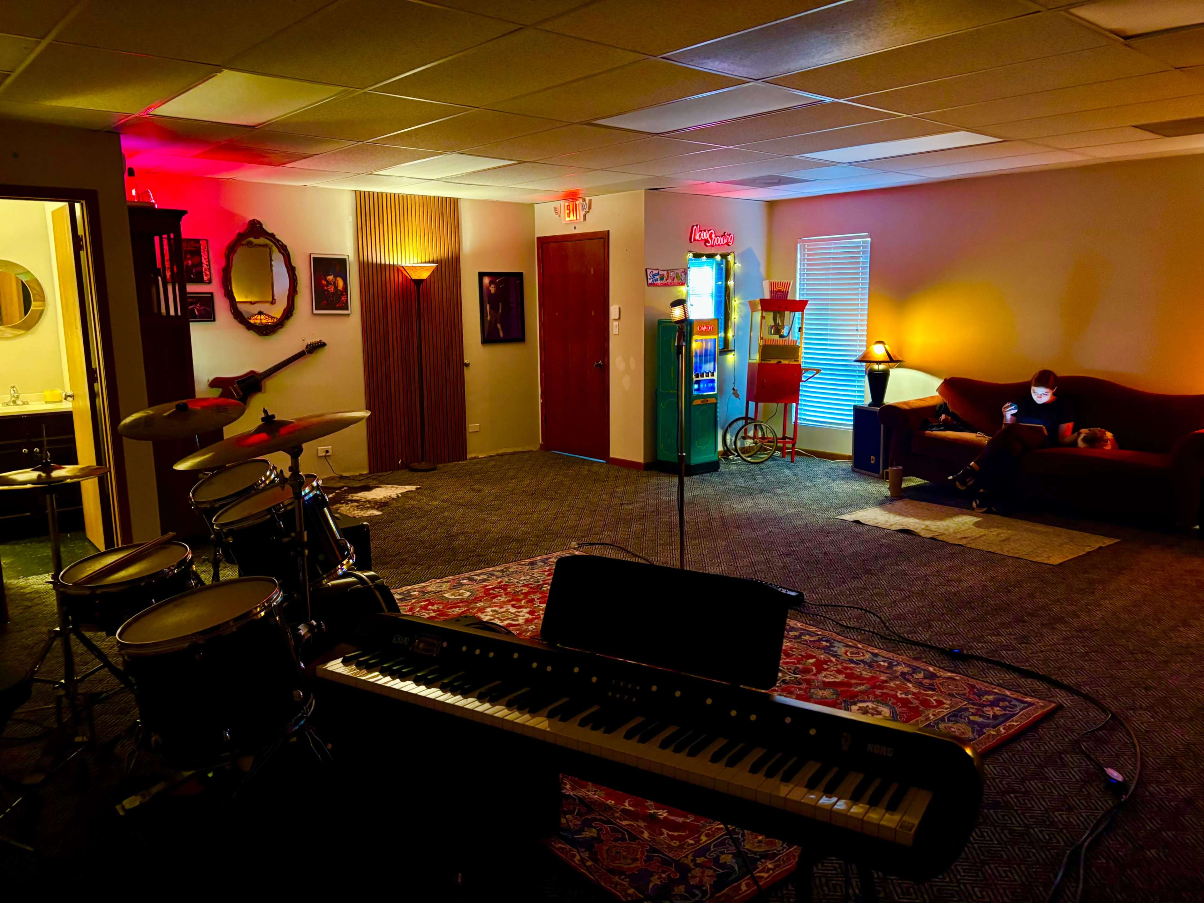 The image shows a spacious music rehearsal room with a drum set and keyboard in the foreground, illuminated by colorful lighting, and a sofa in the background near a vintage arcade machine.