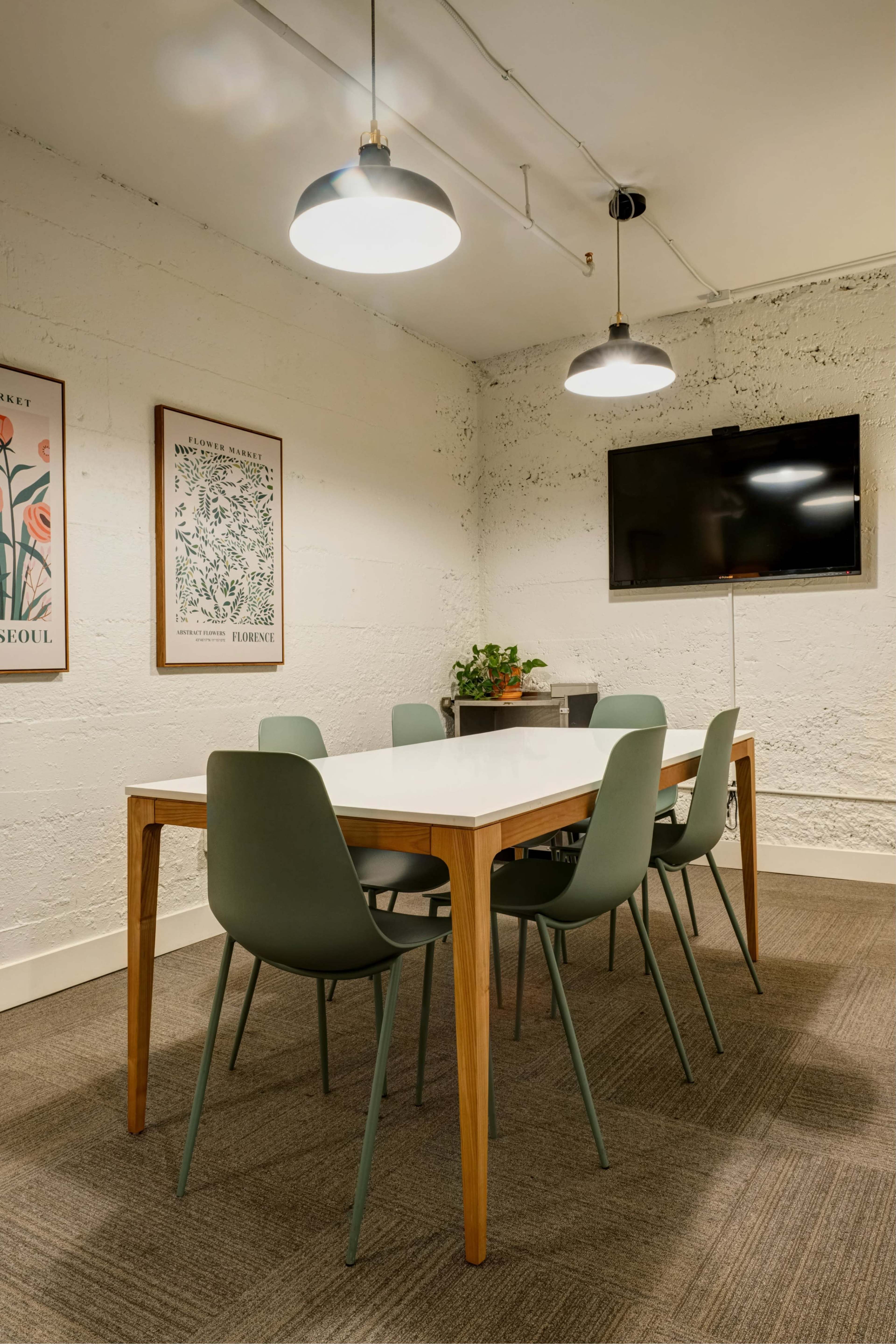 A conference room features a white table surrounded by six green chairs, with a television mounted on the wall and two framed artworks.