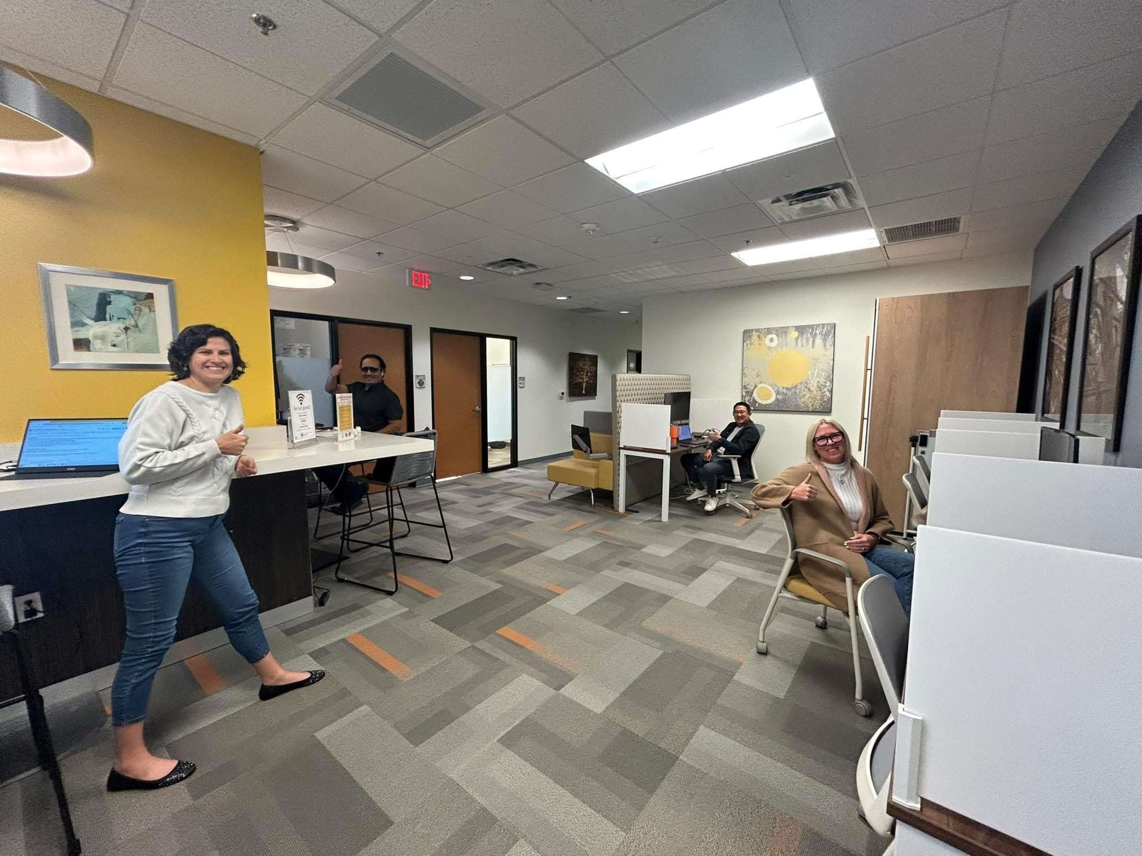 The image shows a modern office space with several people seated and working, along with a person standing in the foreground giving a thumbs-up.