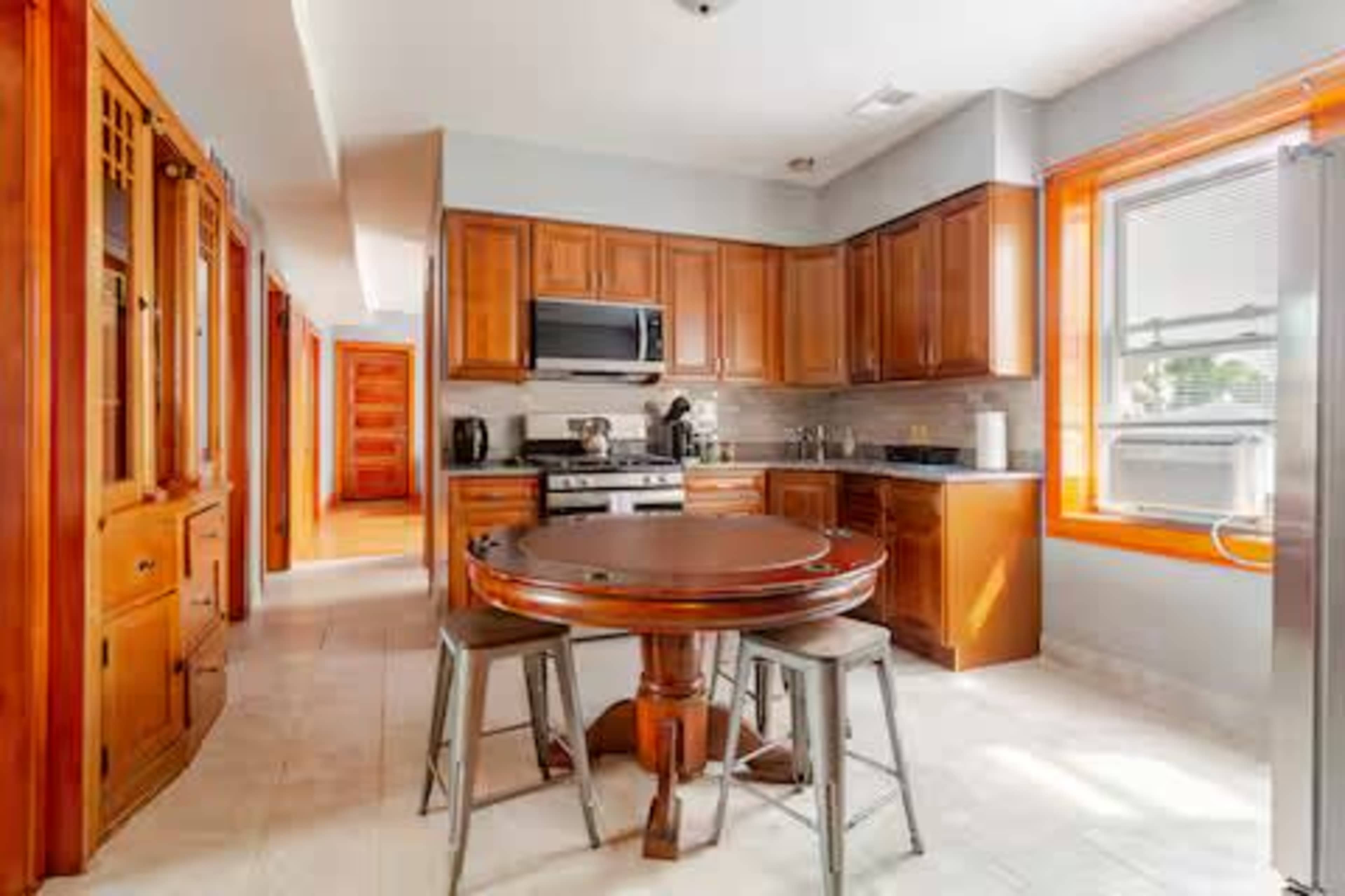 The image shows a modern kitchen featuring wooden cabinetry, stainless steel appliances, and a round dining table with two stools.