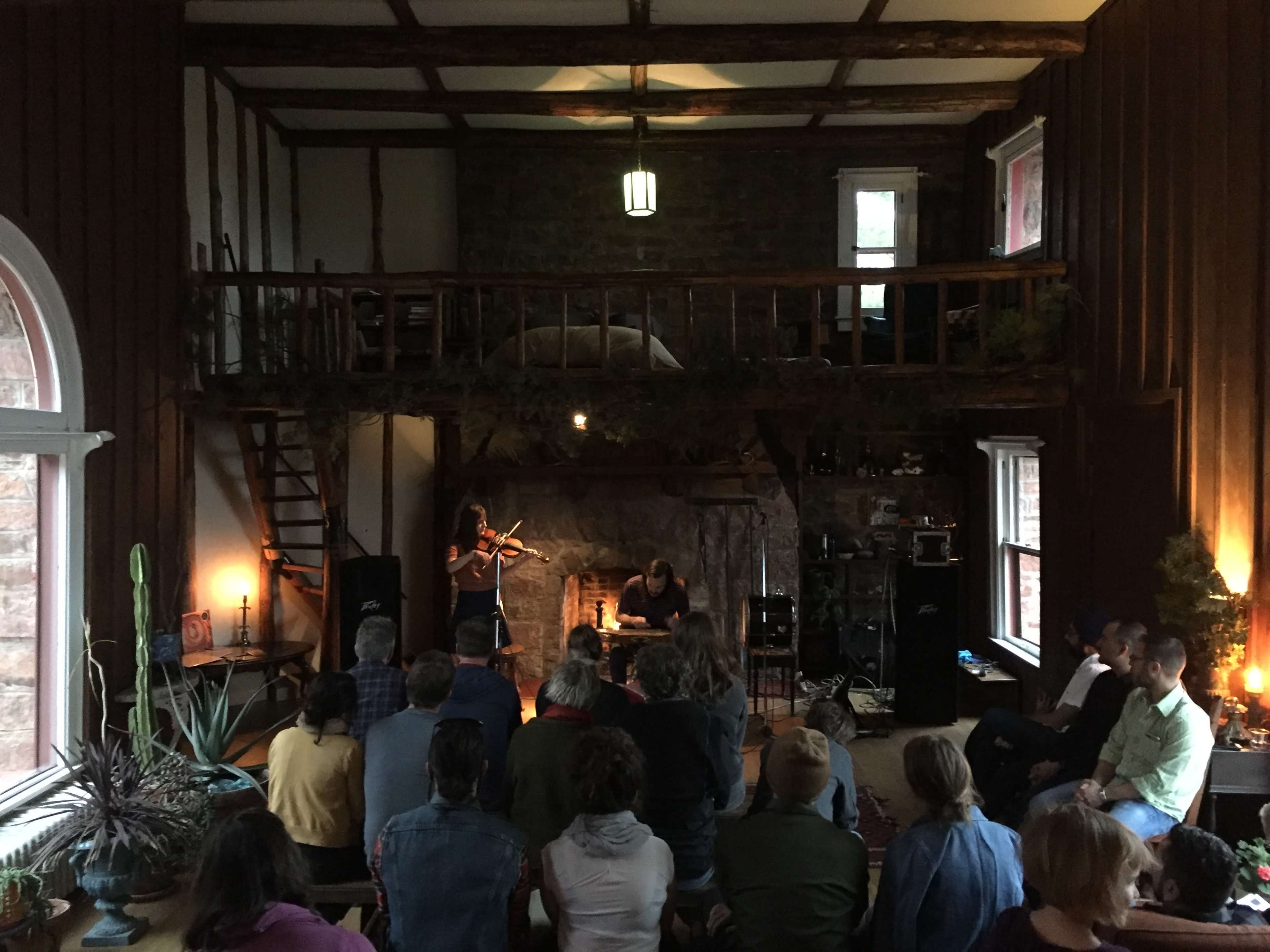 A group of people sit in a cozy room with a stone fireplace, watching a performer play the violin while another musician sits nearby.