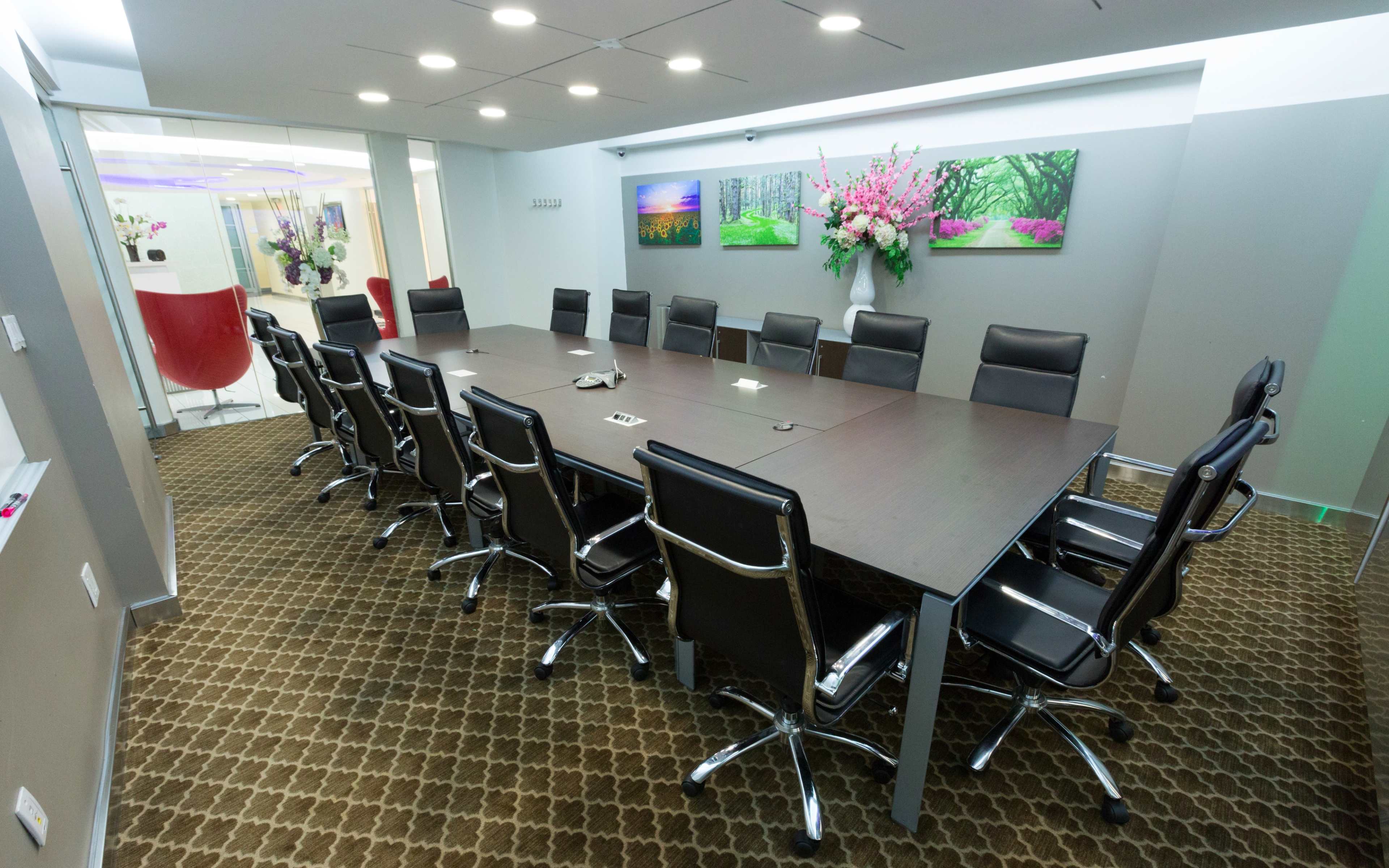 A long conference table with black chairs is set up in a modern meeting room, adorned with floral artwork on the walls.