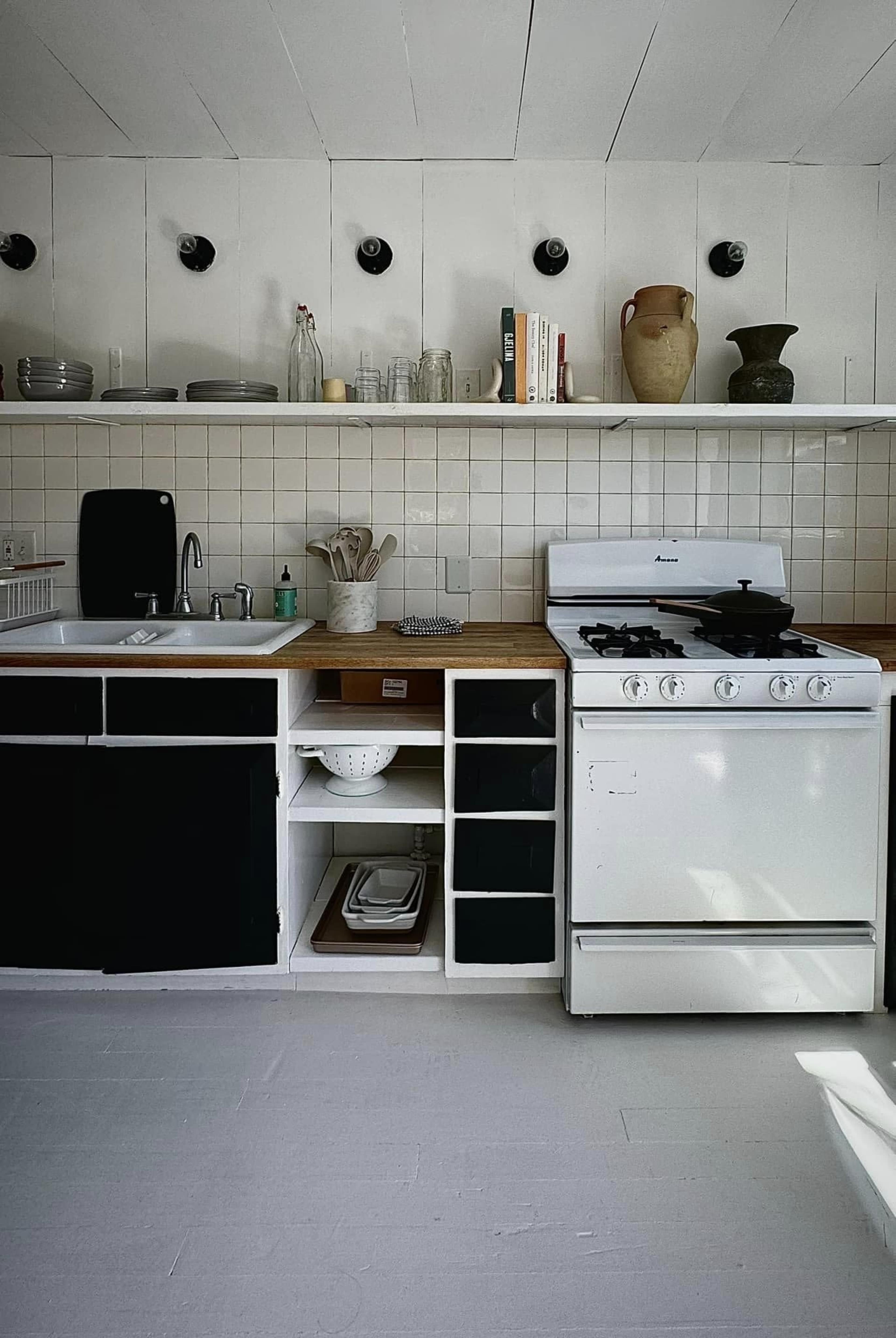 The image shows a kitchen with white walls, wooden countertops, a stainless steel sink, and a white gas stove, along with shelves displaying various kitchenware and decorative items.
