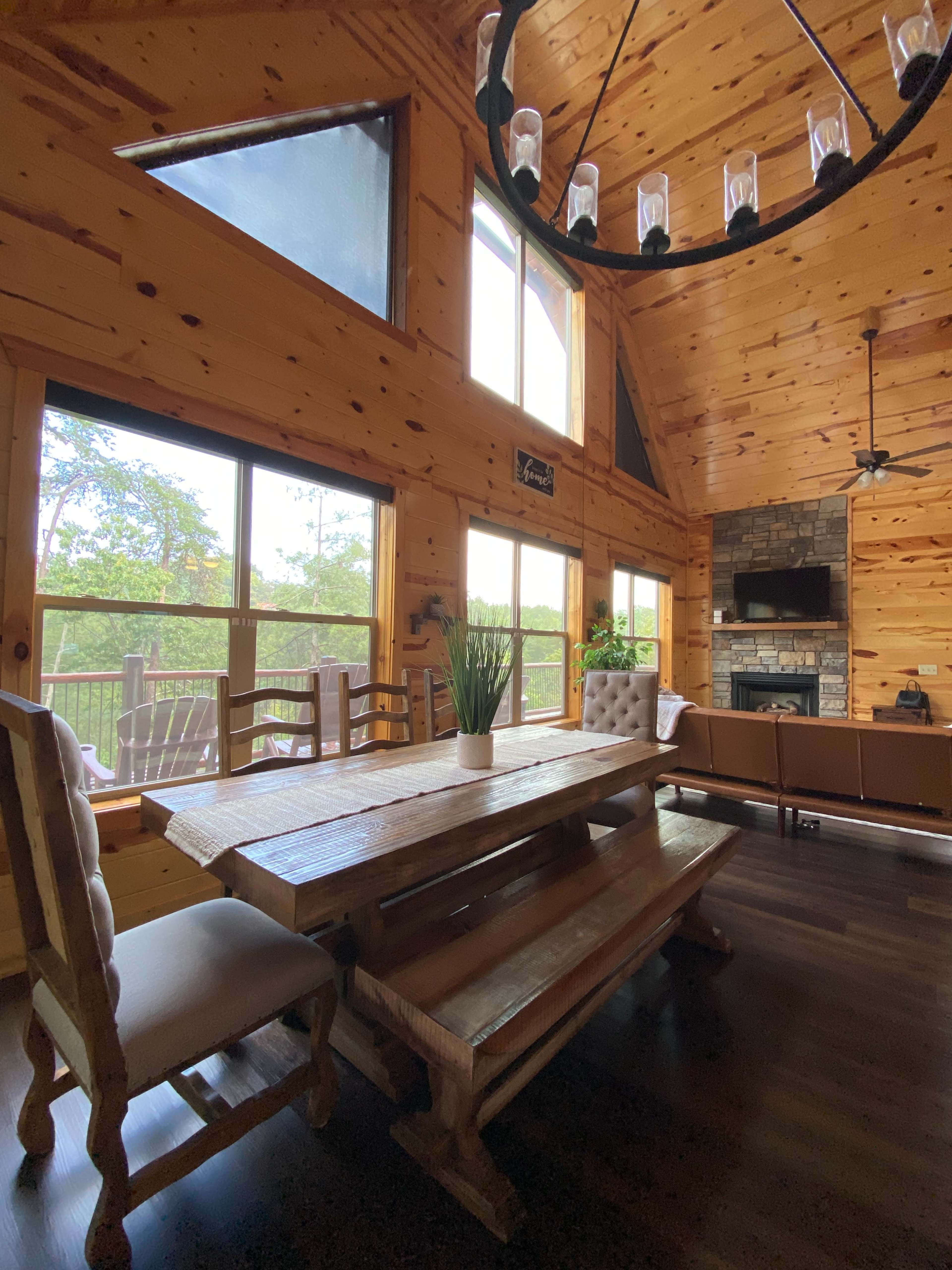 The image shows a wooden dining table with benches in a rustic cabin interior, featuring large windows and a stone fireplace.