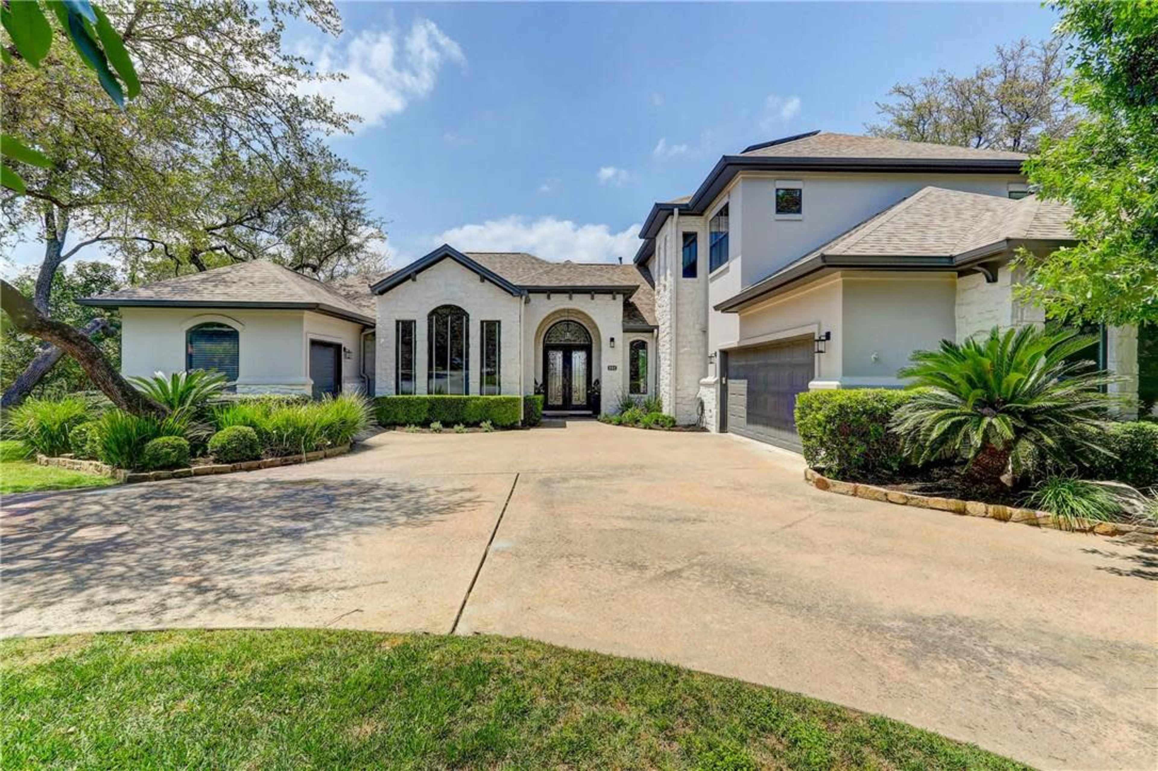 The image shows a modern two-story house with a symmetrical facade, a circular driveway, and landscaped greenery on either side.