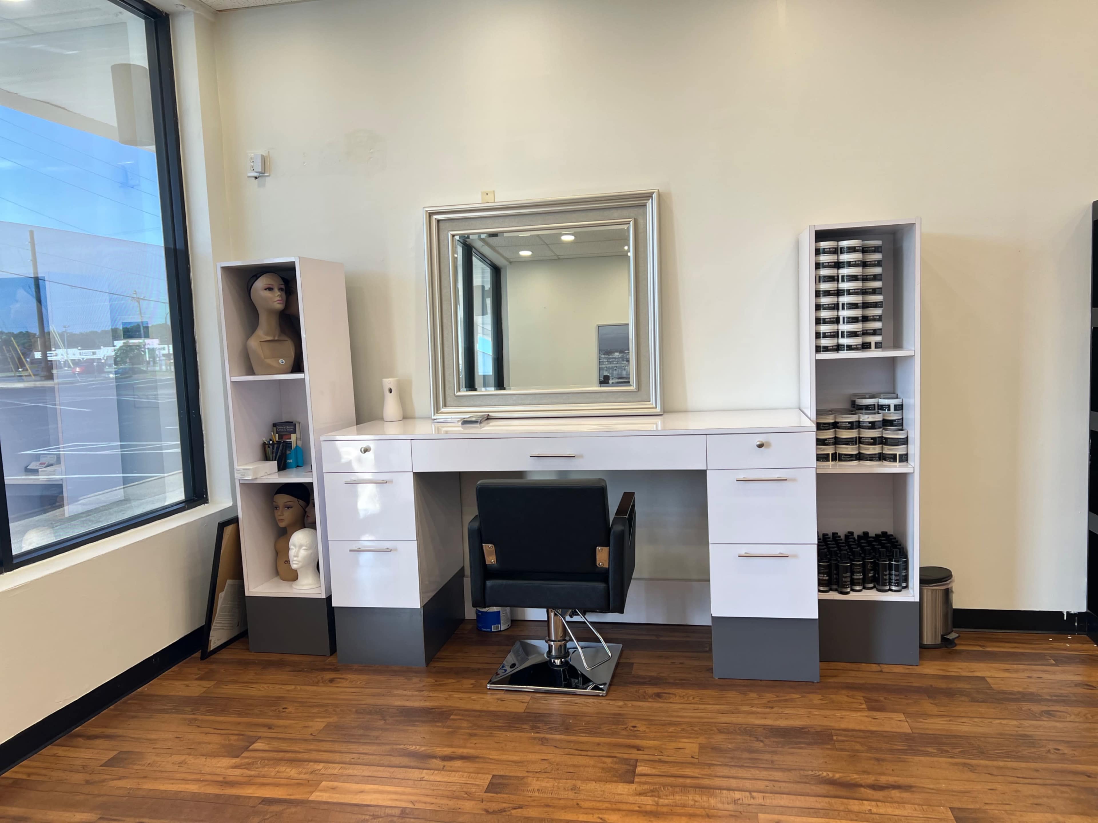 A modern hair salon station with a large mirror, a black salon chair, and shelves containing hair products and mannequin heads.