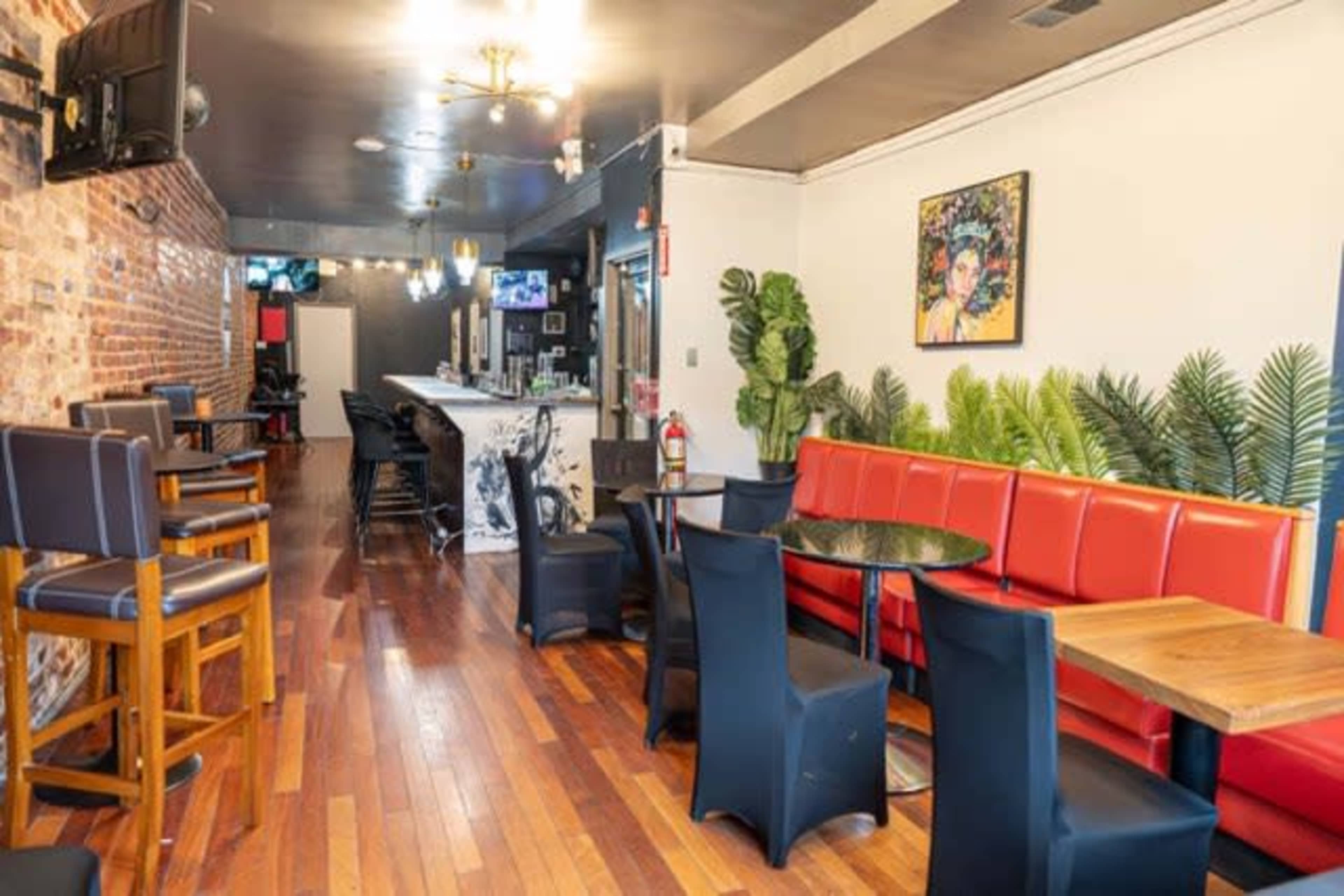 The image shows a cozy bar area with wooden flooring, a red cushioned seating space, black chairs, a bar counter, and decorative plants along the walls.