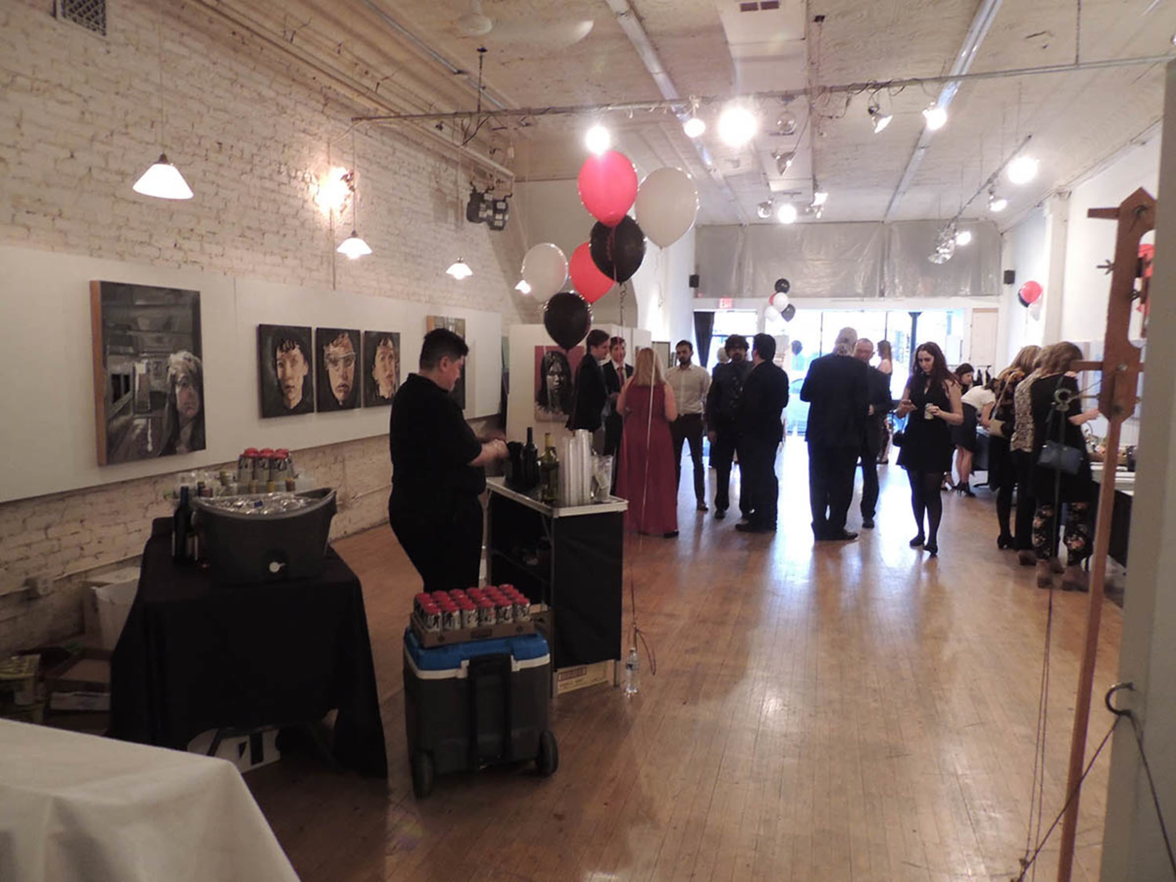 A spacious art gallery with guests mingling, a black table with drinks on one side, and artwork hanging on the walls.