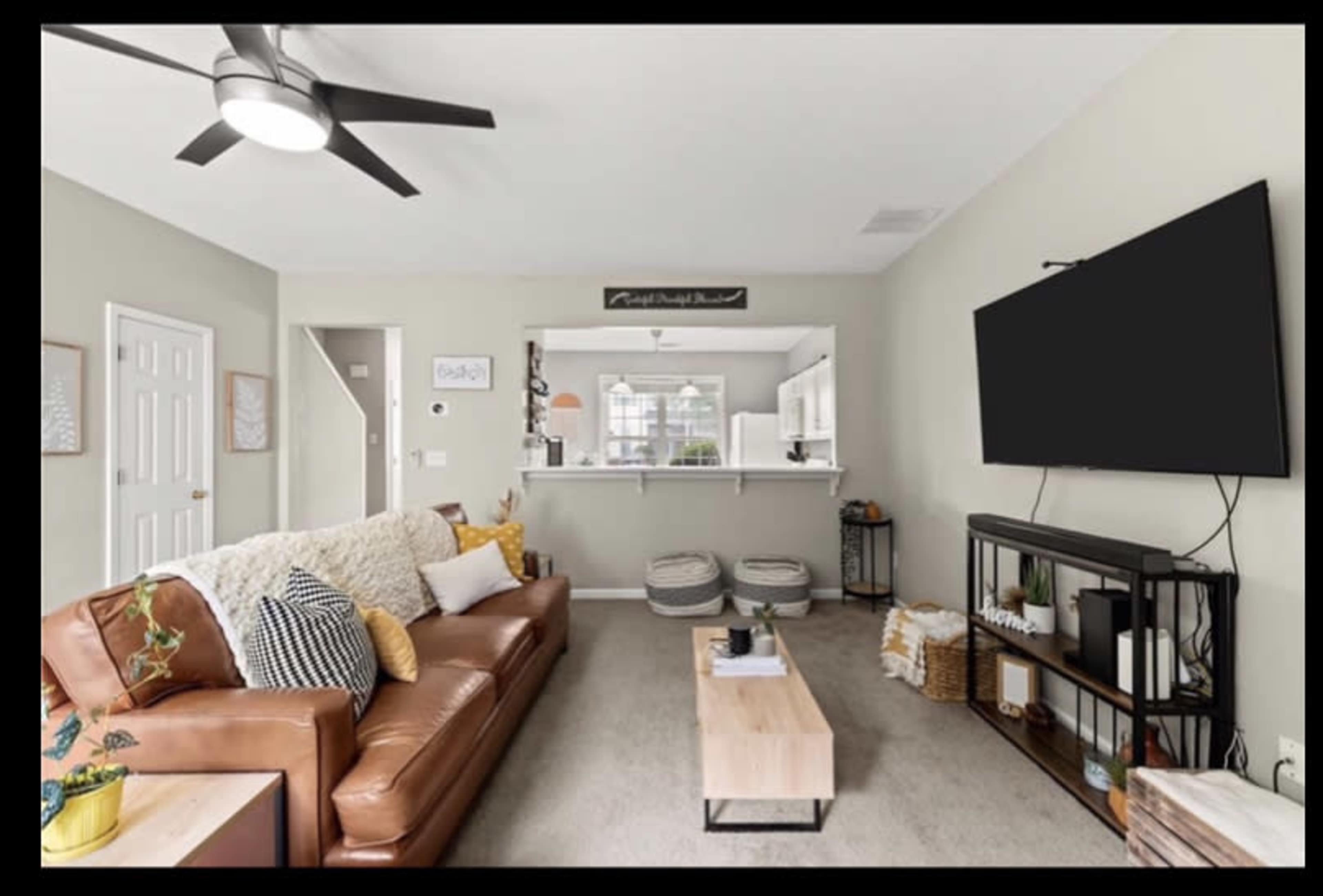 The image shows a modern living room with a brown leather sofa, a wooden coffee table, and a television mounted on a wall, with soft light filtering through the windows.