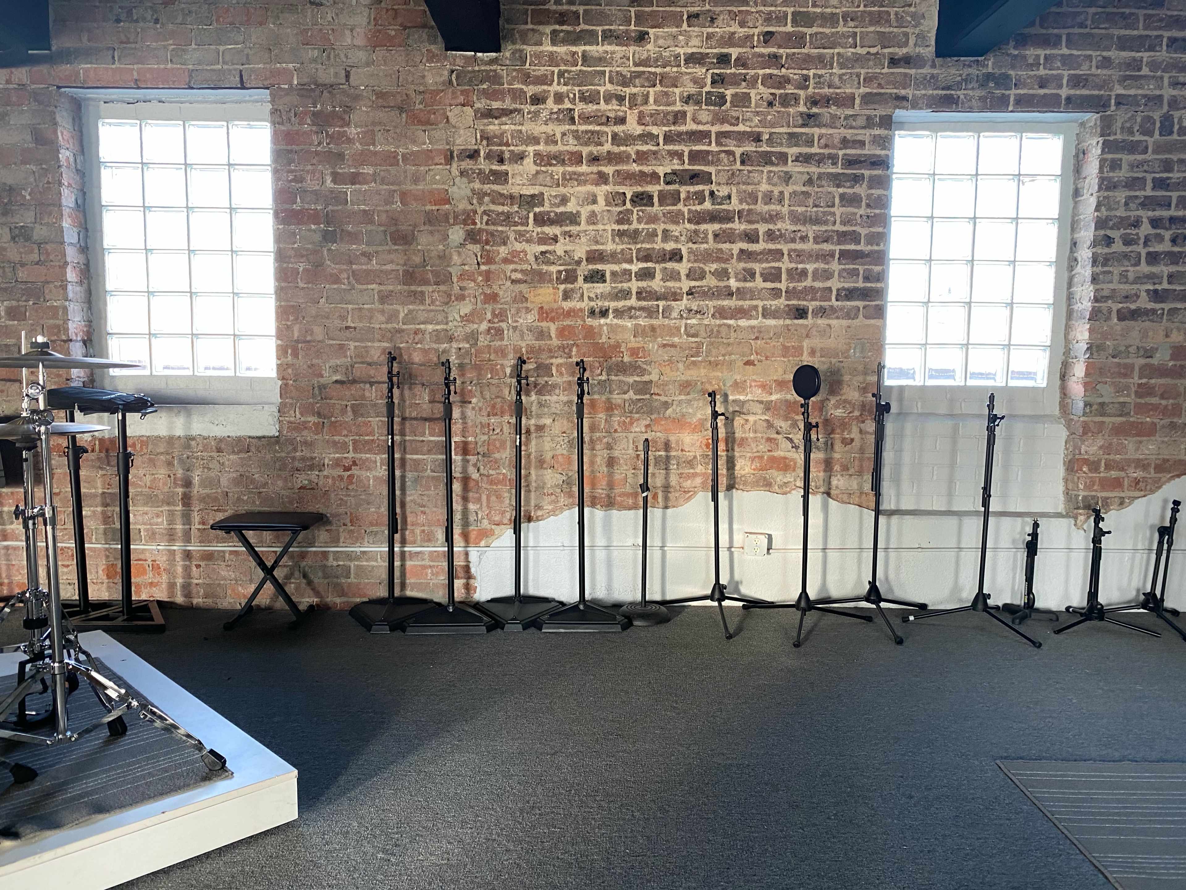 The image shows a brick wall with two windows and several microphone stands lined up against the wall, beside a drum set on a platform.
