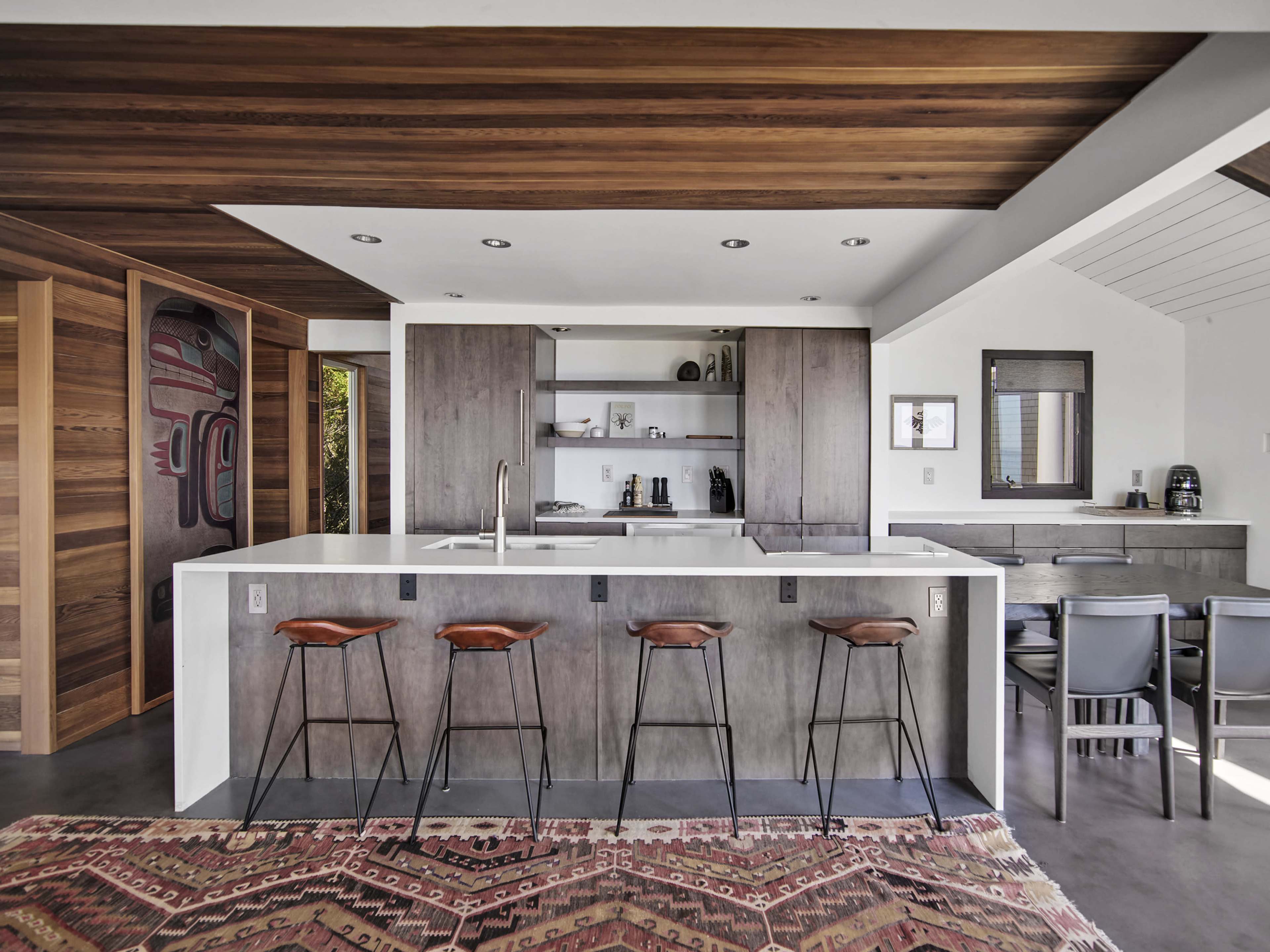 A modern kitchen with a white island, six bar stools, gray cabinetry, and a dining area in the background.