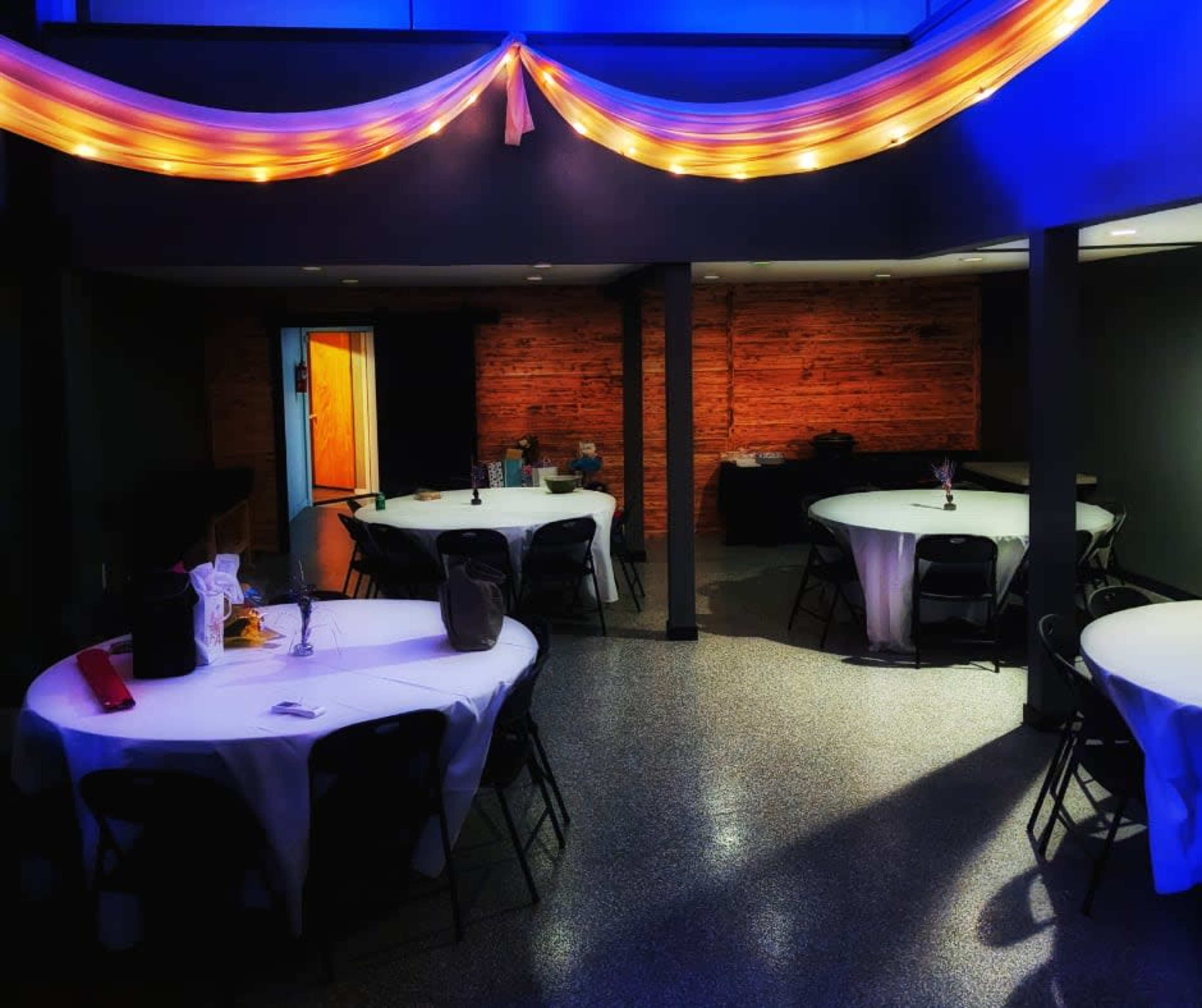 A spacious event venue features circular tables set with white tablecloths, illuminated by colorful lighting and decorated with a flowing fabric drape overhead.