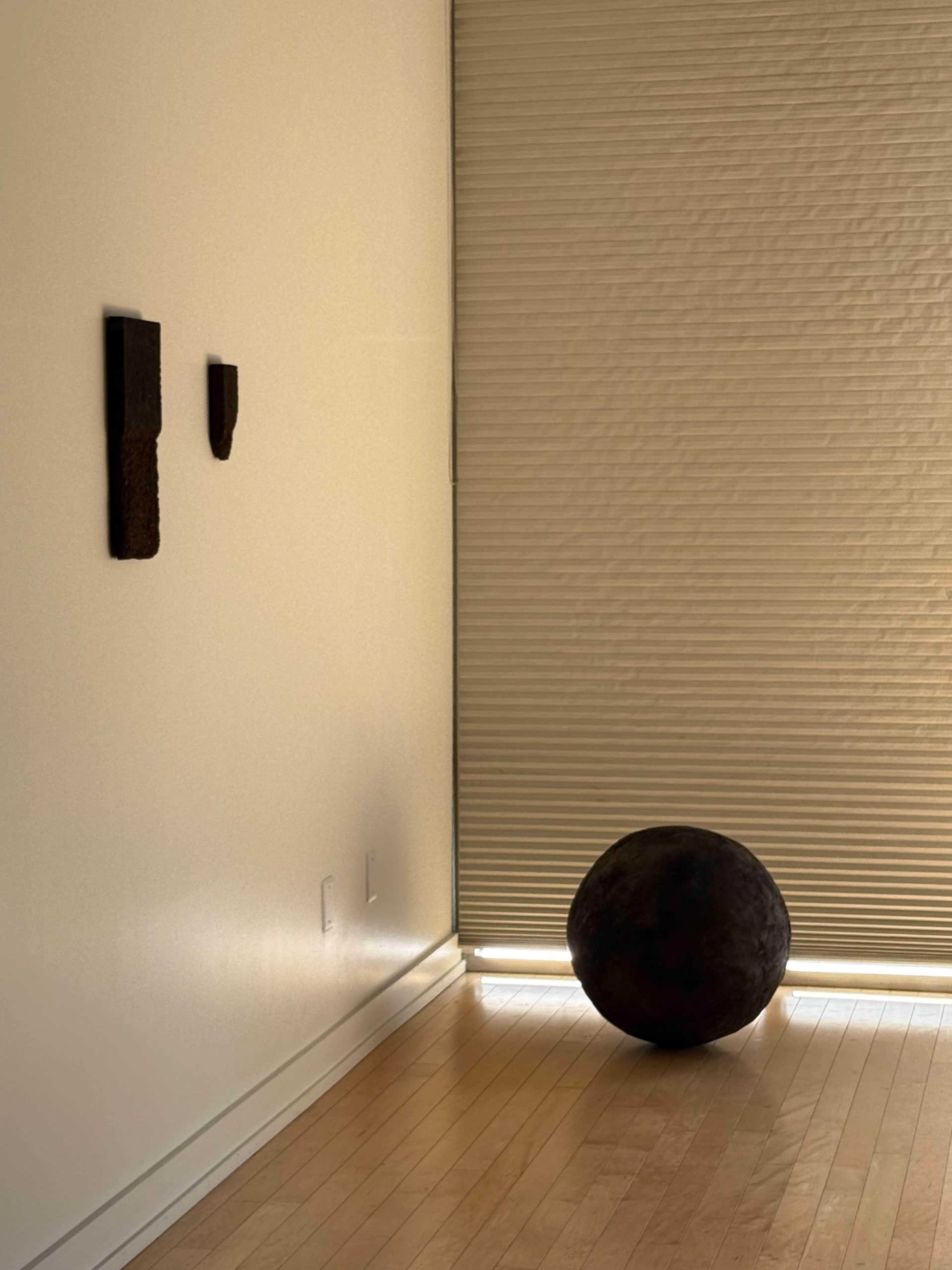 A round dark object sits on a wooden floor in a corner next to a textured wall with two dark pieces attached, while light filters through a closed window covering.