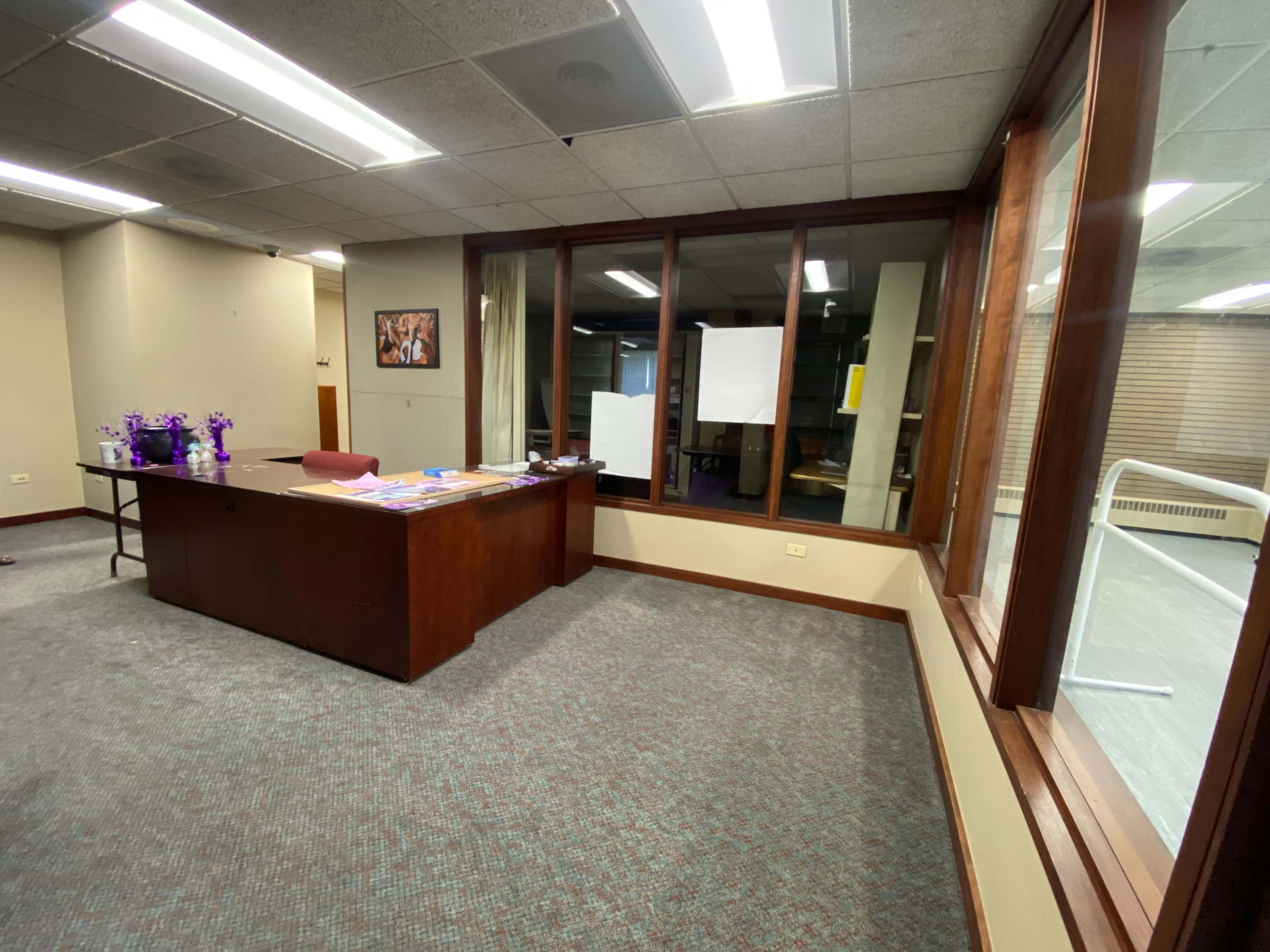 An office space with a wooden desk, scattered papers, and large windows that provide a view of an adjacent area.