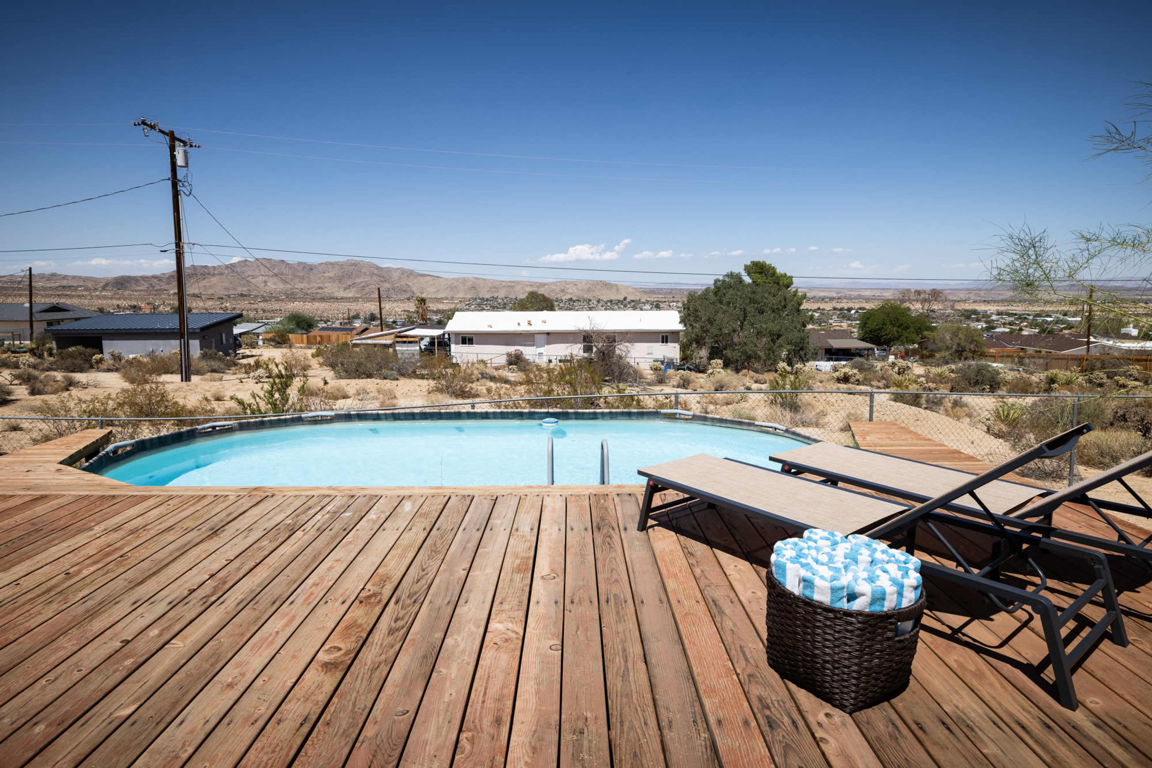 The image shows a wooden deck with a swimming pool and a lounge chair overlooking a desert landscape with distant hills and houses.