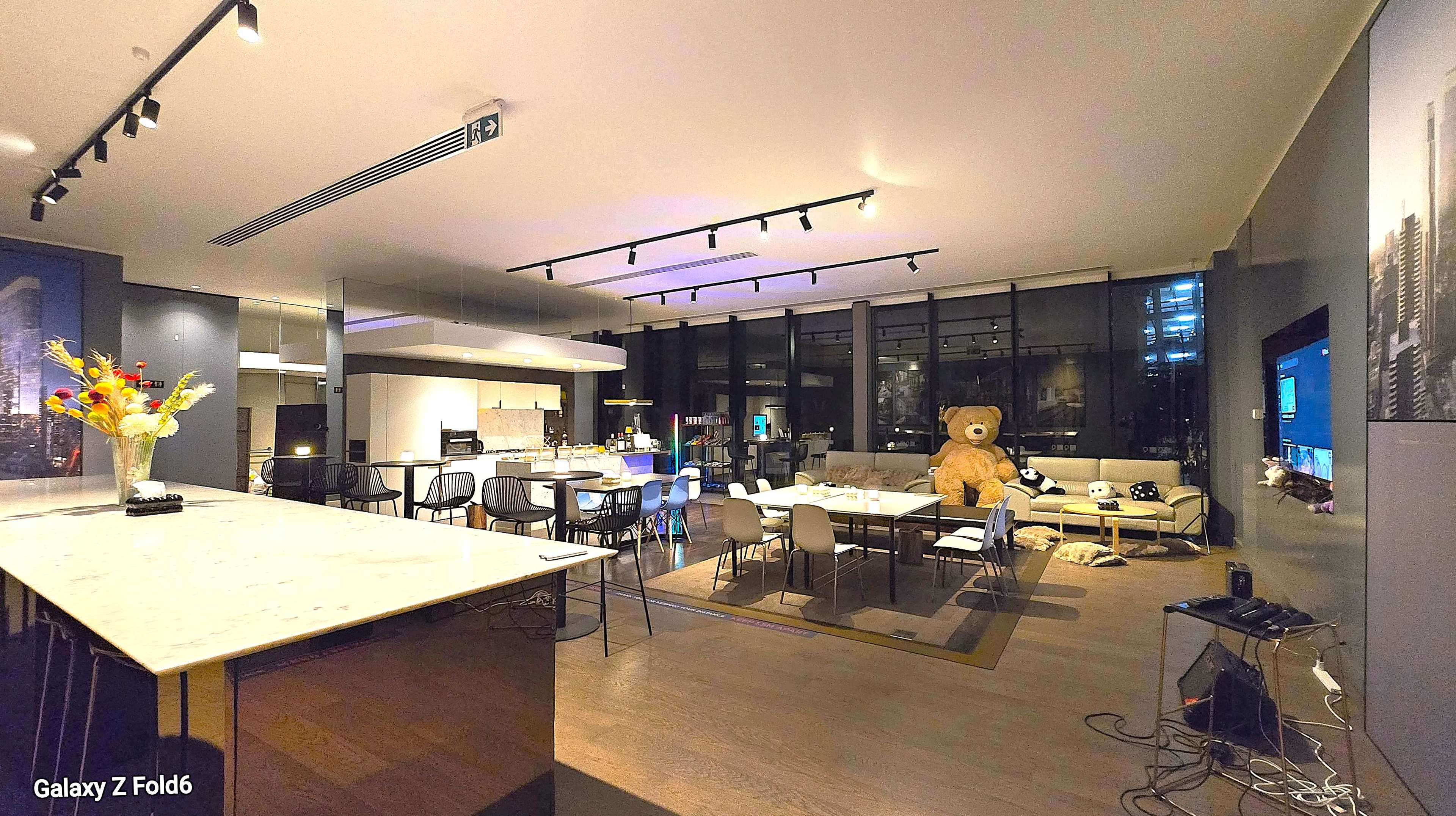 A spacious modern living area features a large kitchen island, dining table, and a bear-shaped stuffed toy in a corner, with large windows providing city views.