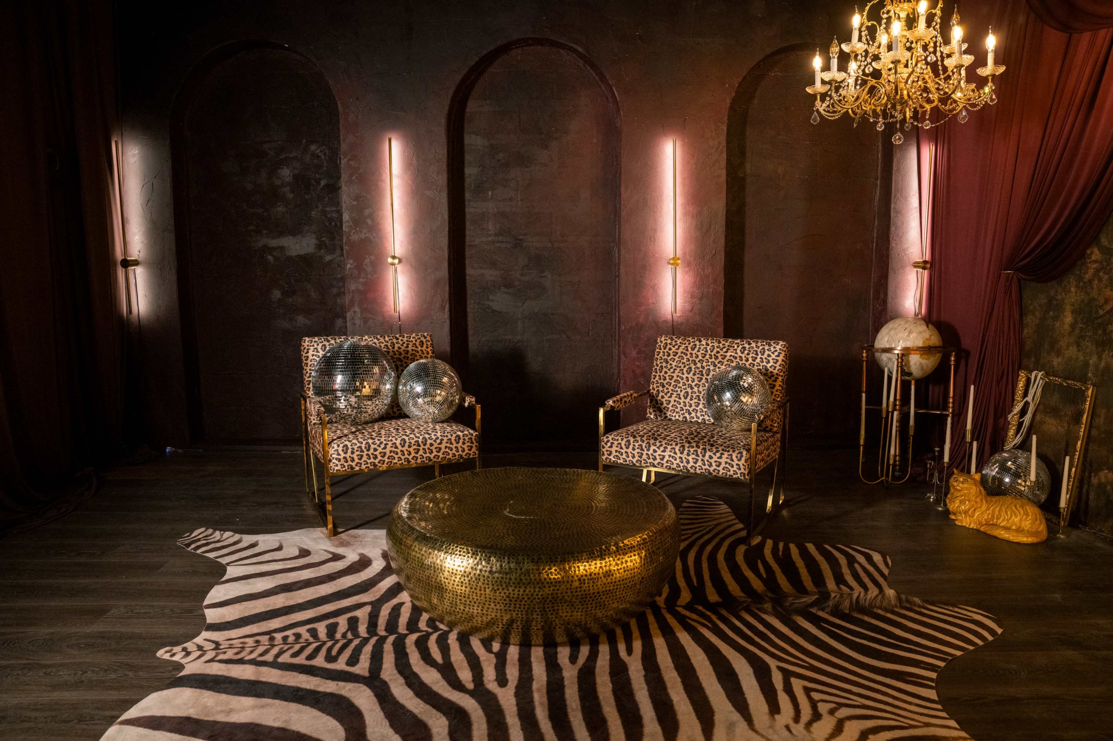 The scene features two leopard-print chairs facing a round, gold coffee table on a zebra-striped rug, with a chandelier hanging above in a dimly lit room.