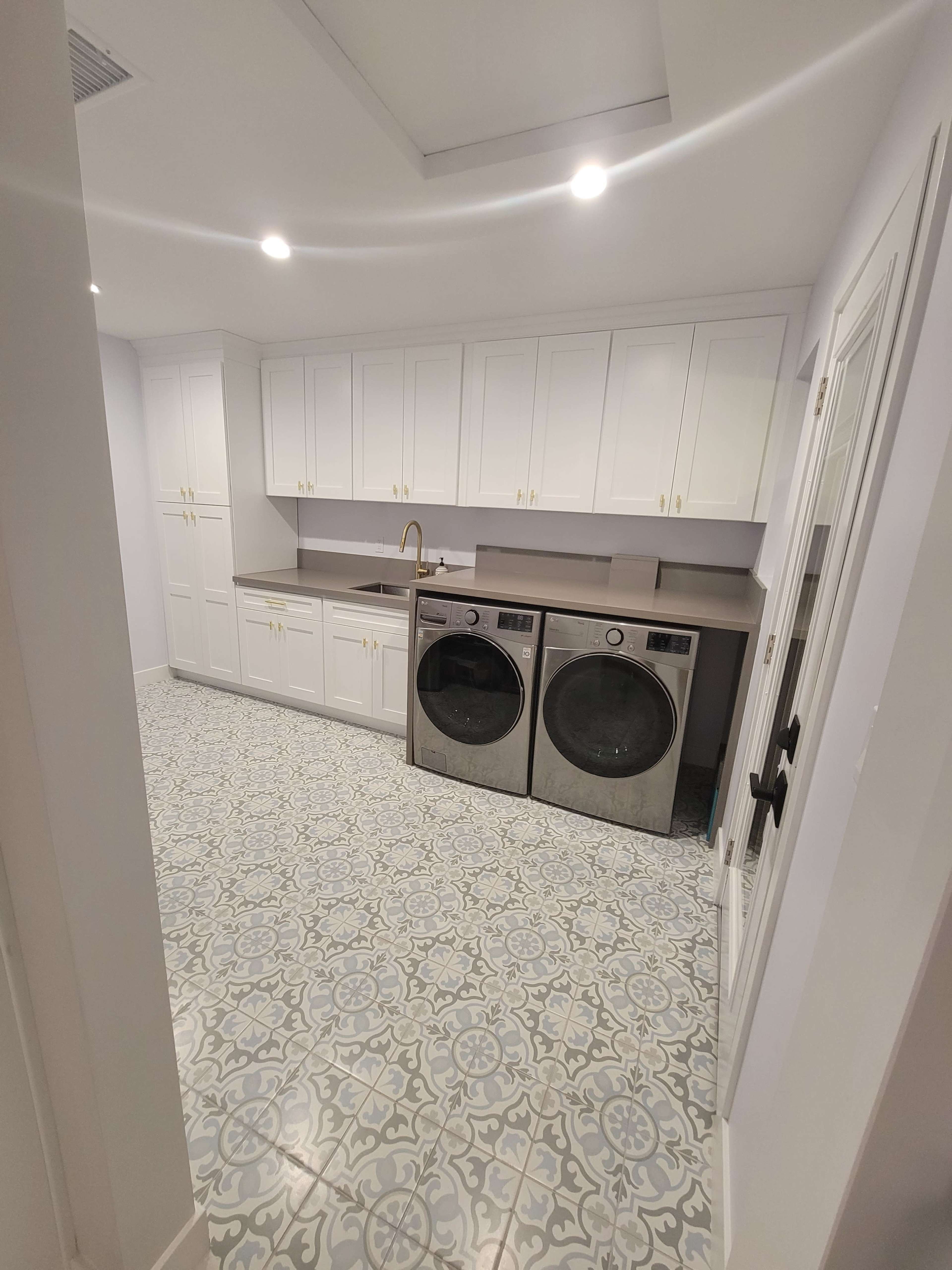 A laundry room features white cabinetry, a gray countertop, and front-loading washing machines on patterned floor tiles.