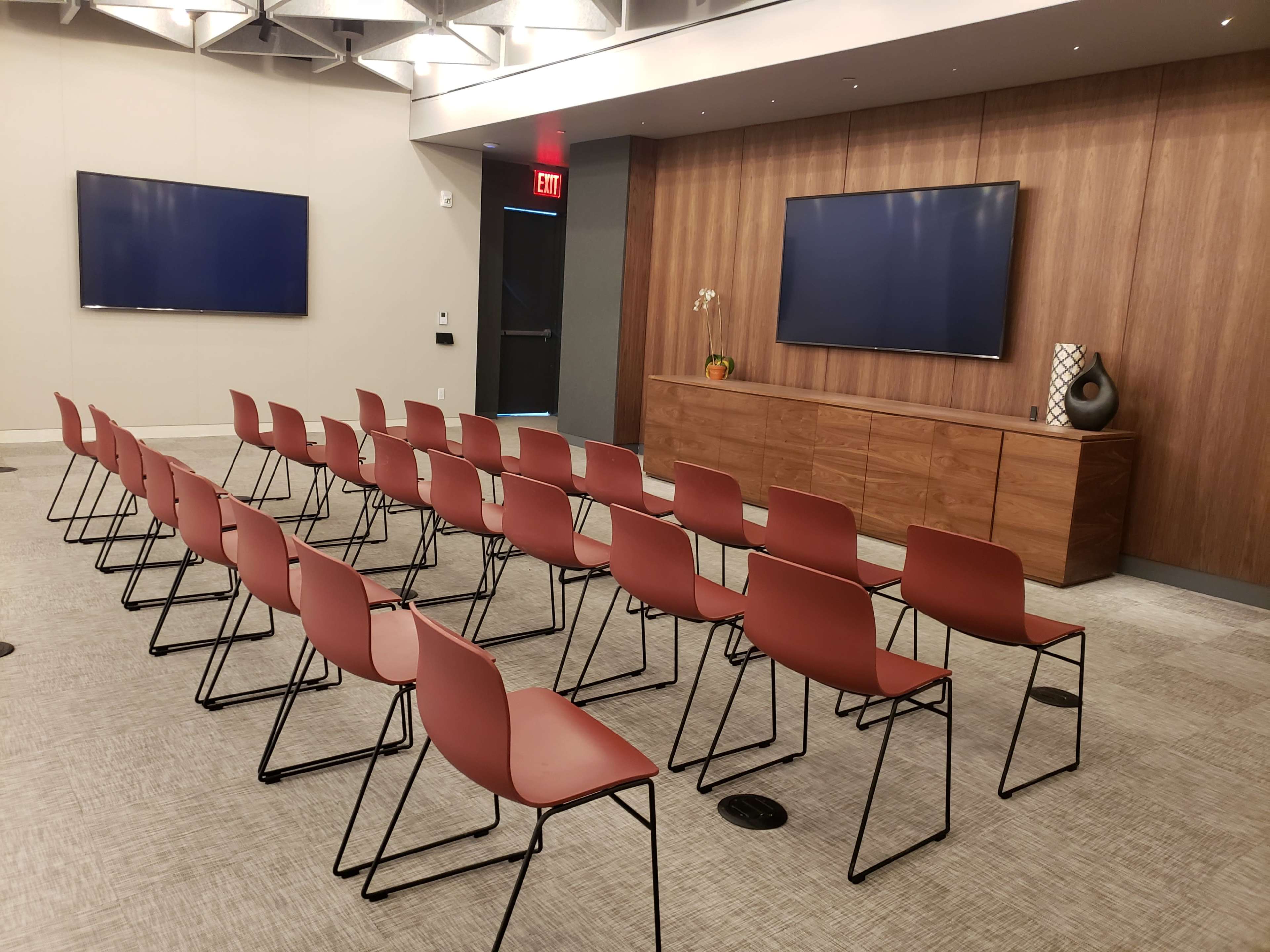A meeting room with rows of red chairs facing two wall-mounted screens and a wooden cabinet.