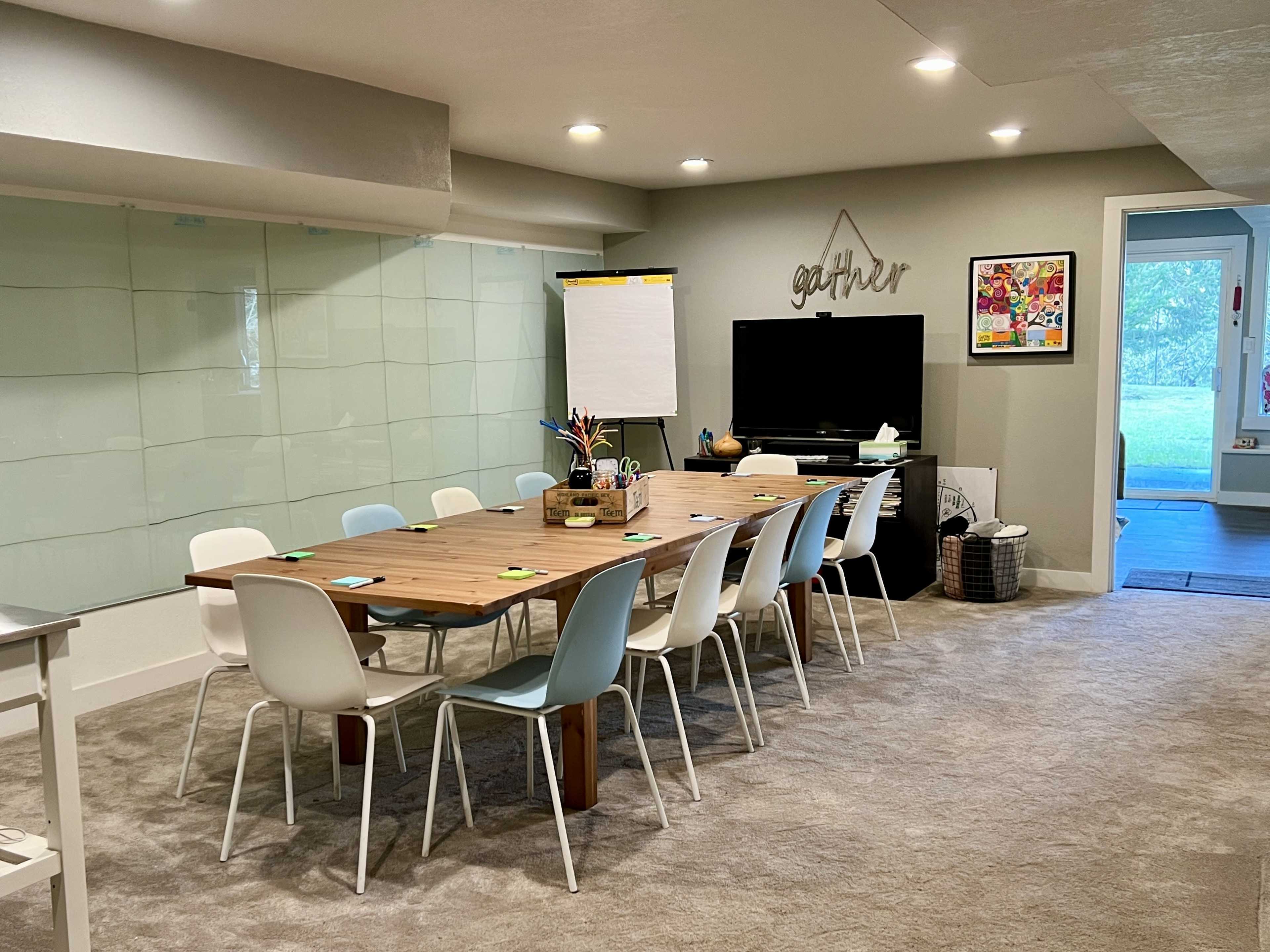 A long wooden table with white and light blue chairs is set up in a well-lit meeting room featuring a glass wall, a TV, and various supplies on the table.