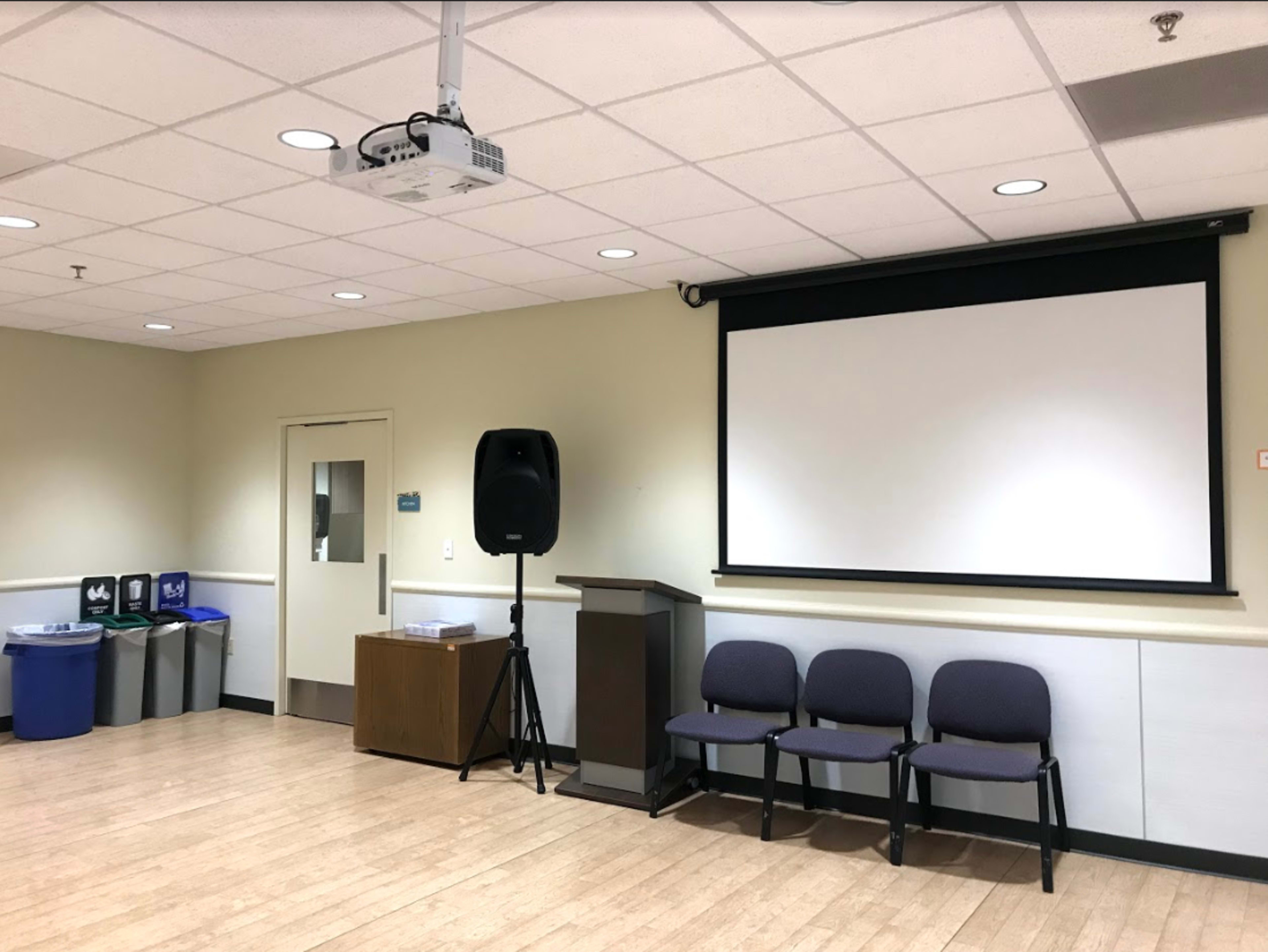 The image shows a room set up for a presentation, featuring a projector, a large screen, a podium, three chairs, and recycling bins in the corner.