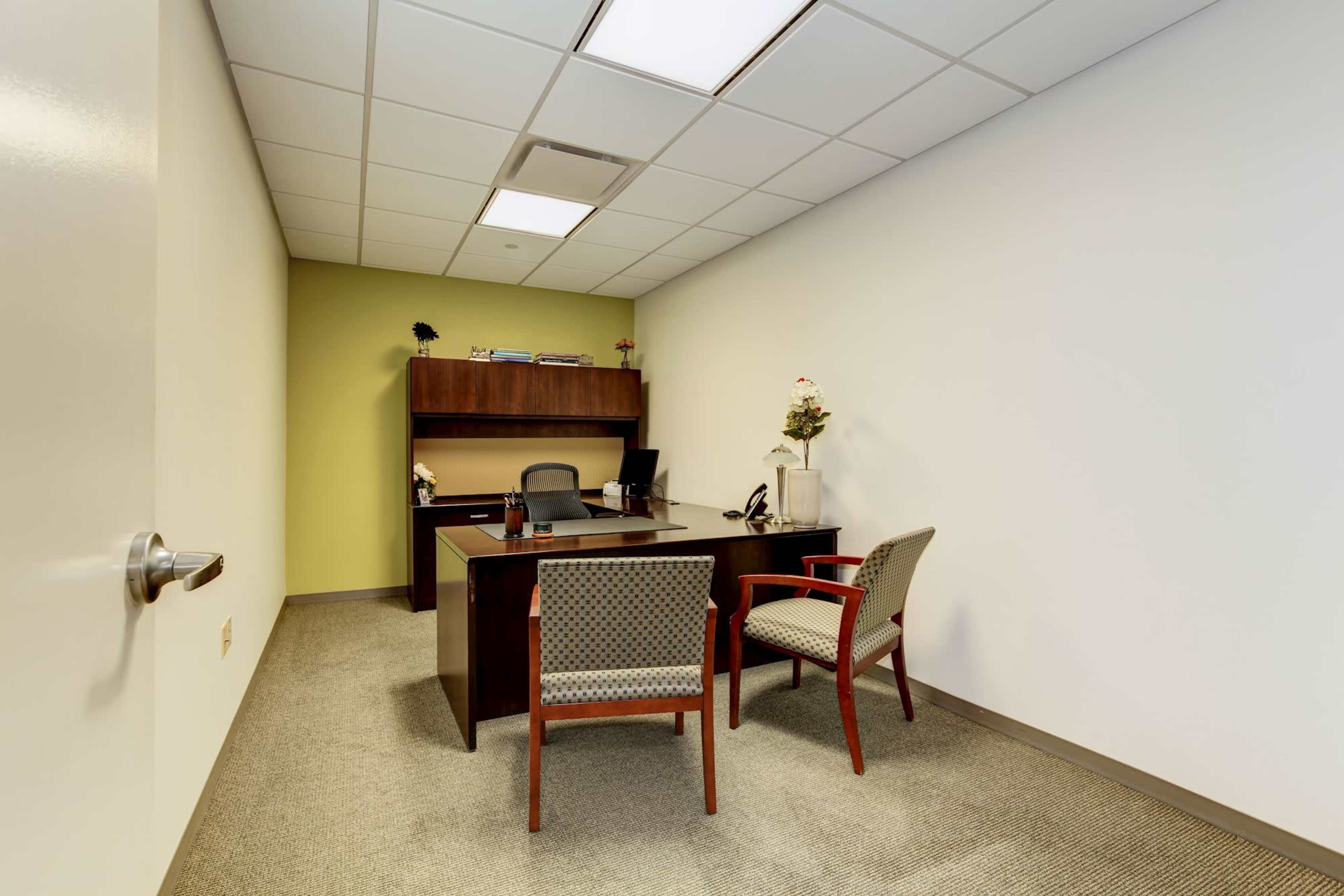 The image shows a small office with a desk, two chairs, and a shelf against a wall.
