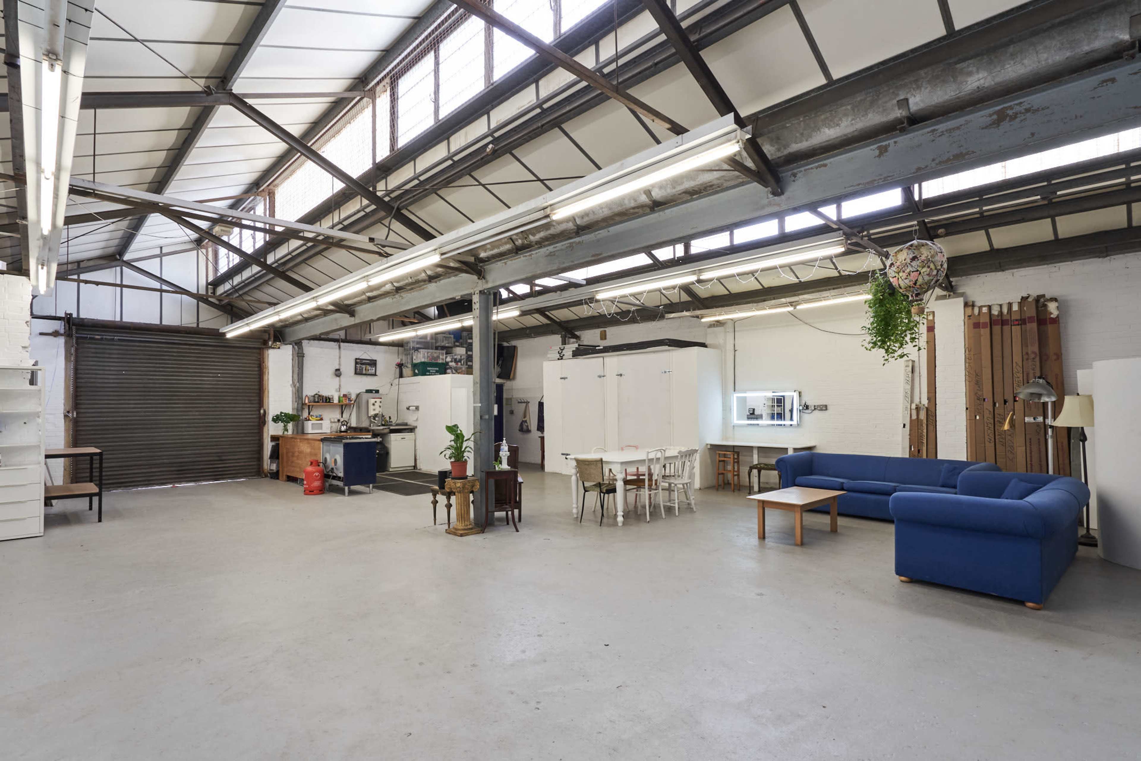 A spacious, industrial-style room featuring a blue sofa, a wooden coffee table, a dining area with white chairs, and large windows overhead.