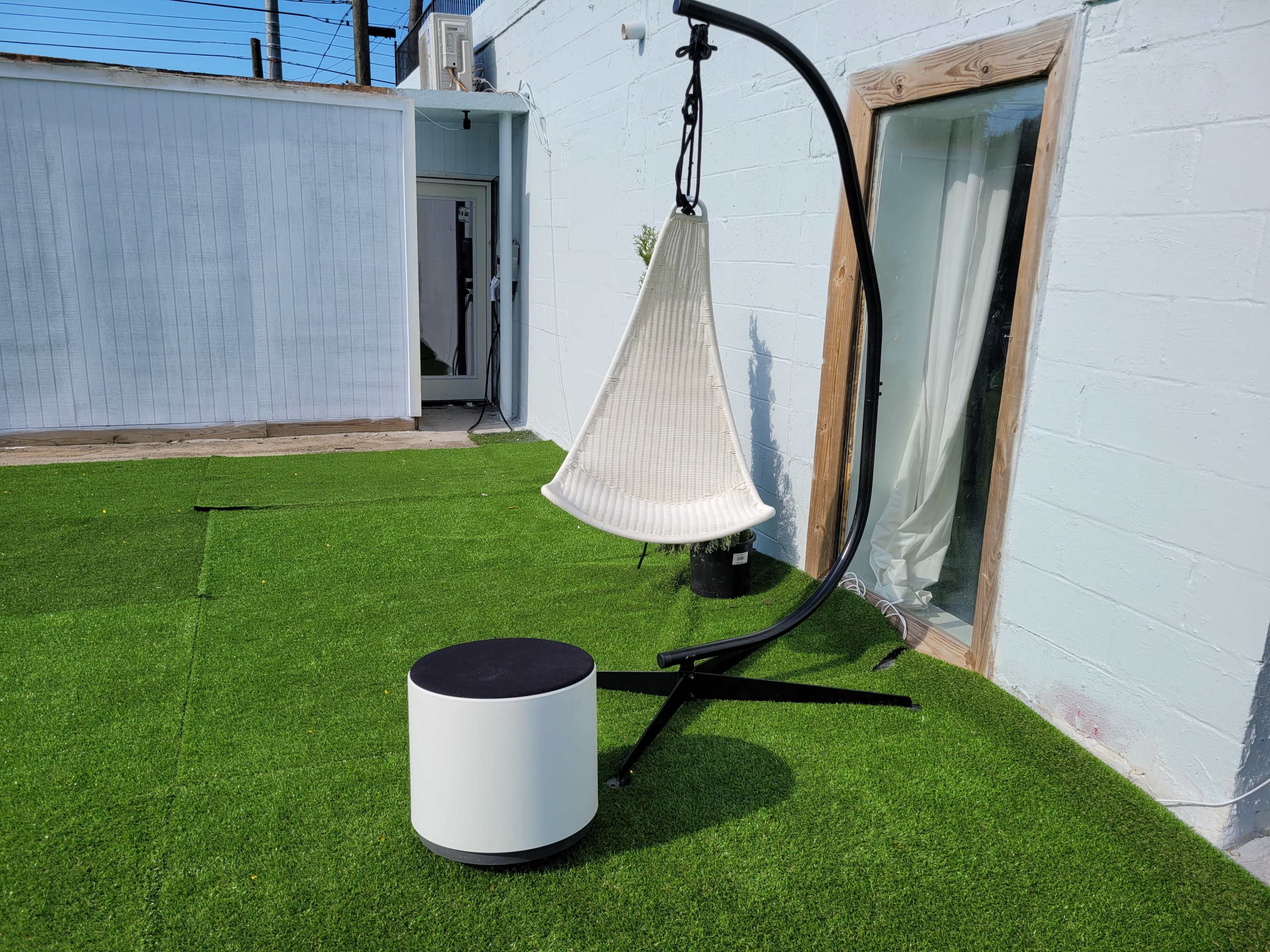 A suspended chair is attached to a black stand on a grass-like surface next to a white wall and a doorway.