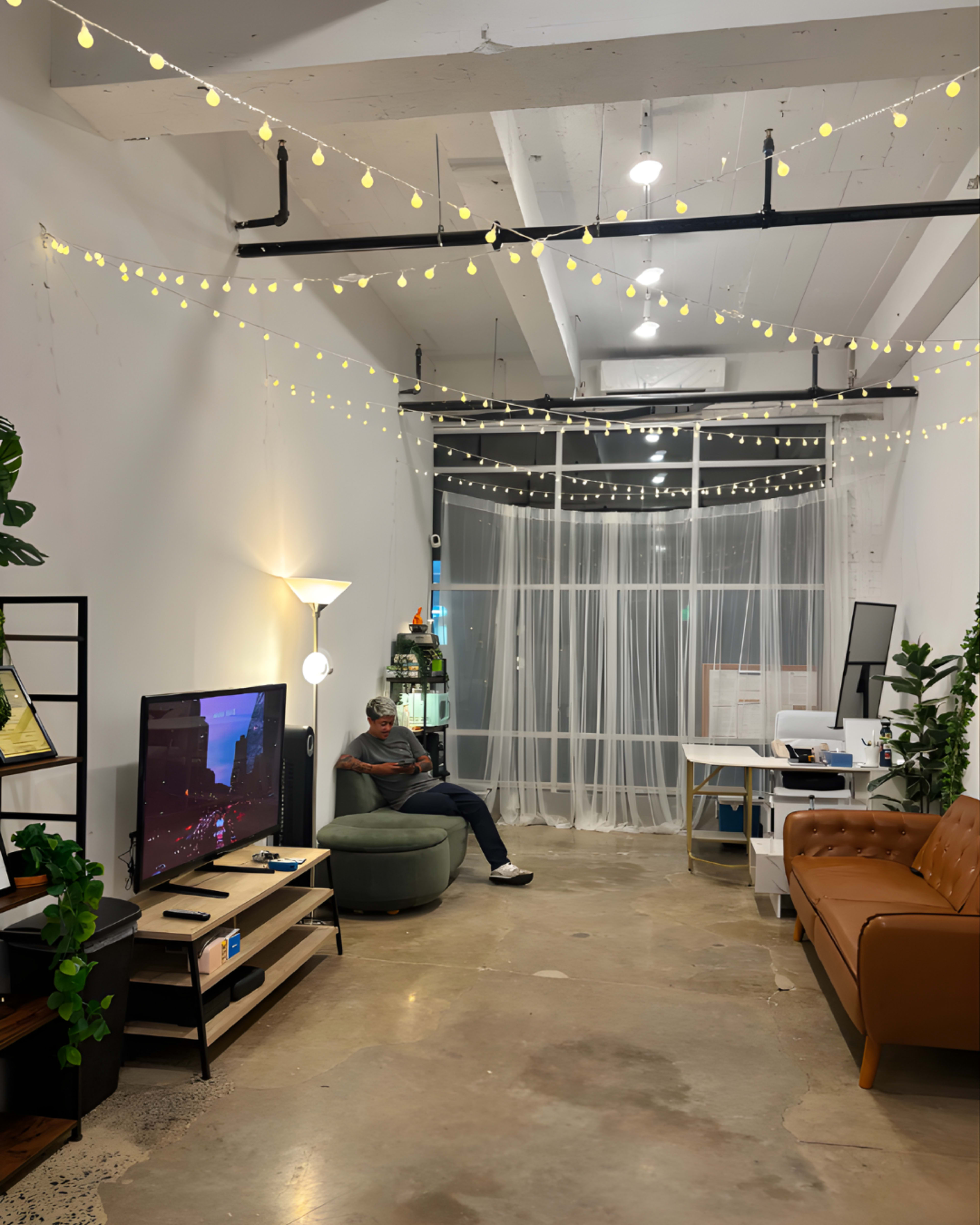The image shows a modern office space with a couch, a TV on a stand, a person sitting on a round chair, and decorative string lights overhead.