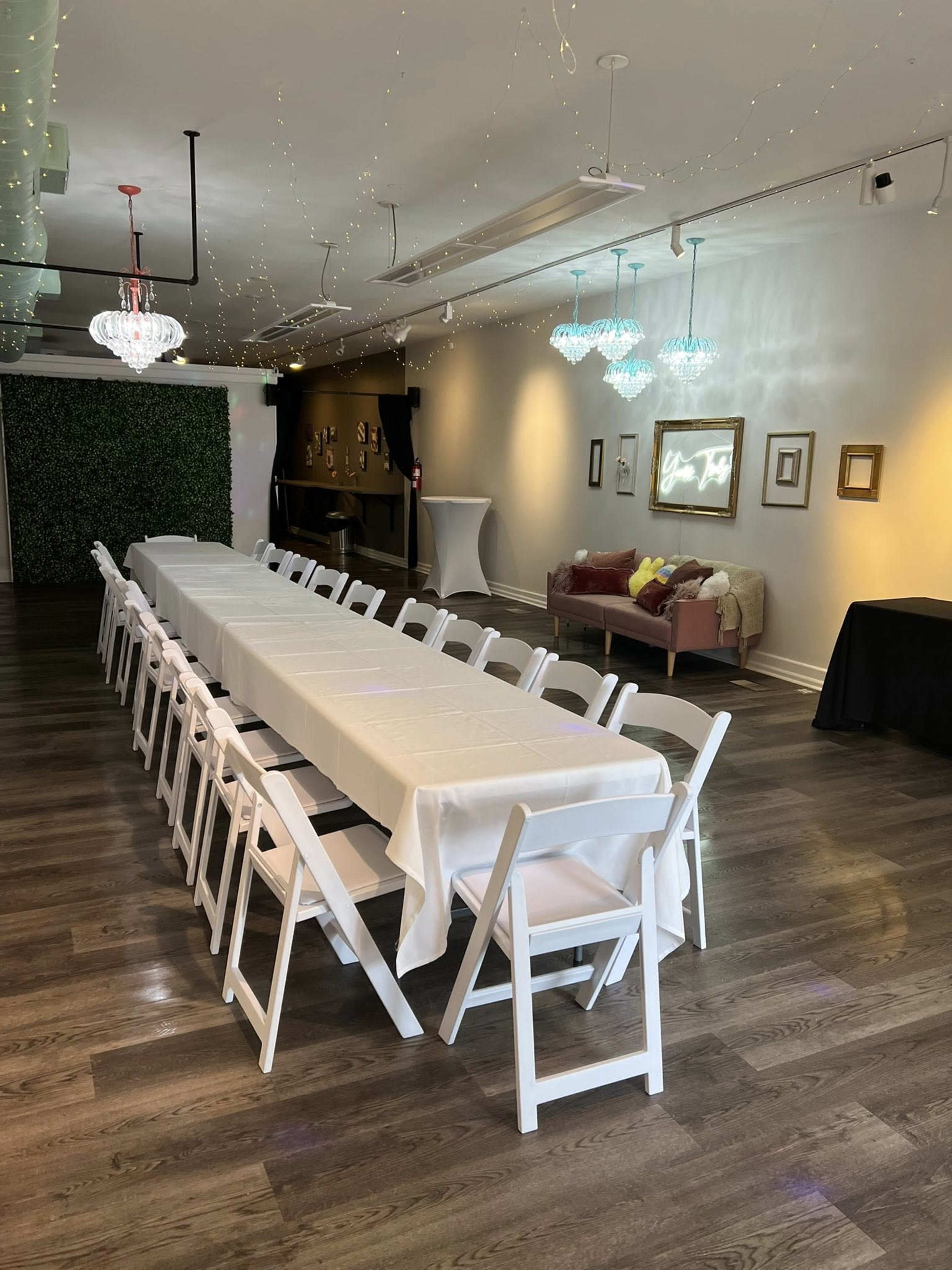 A long, neatly arranged table with white tablecloths and folding chairs is set up in a well-lit event space, featuring decorative lighting and a cozy seating area.