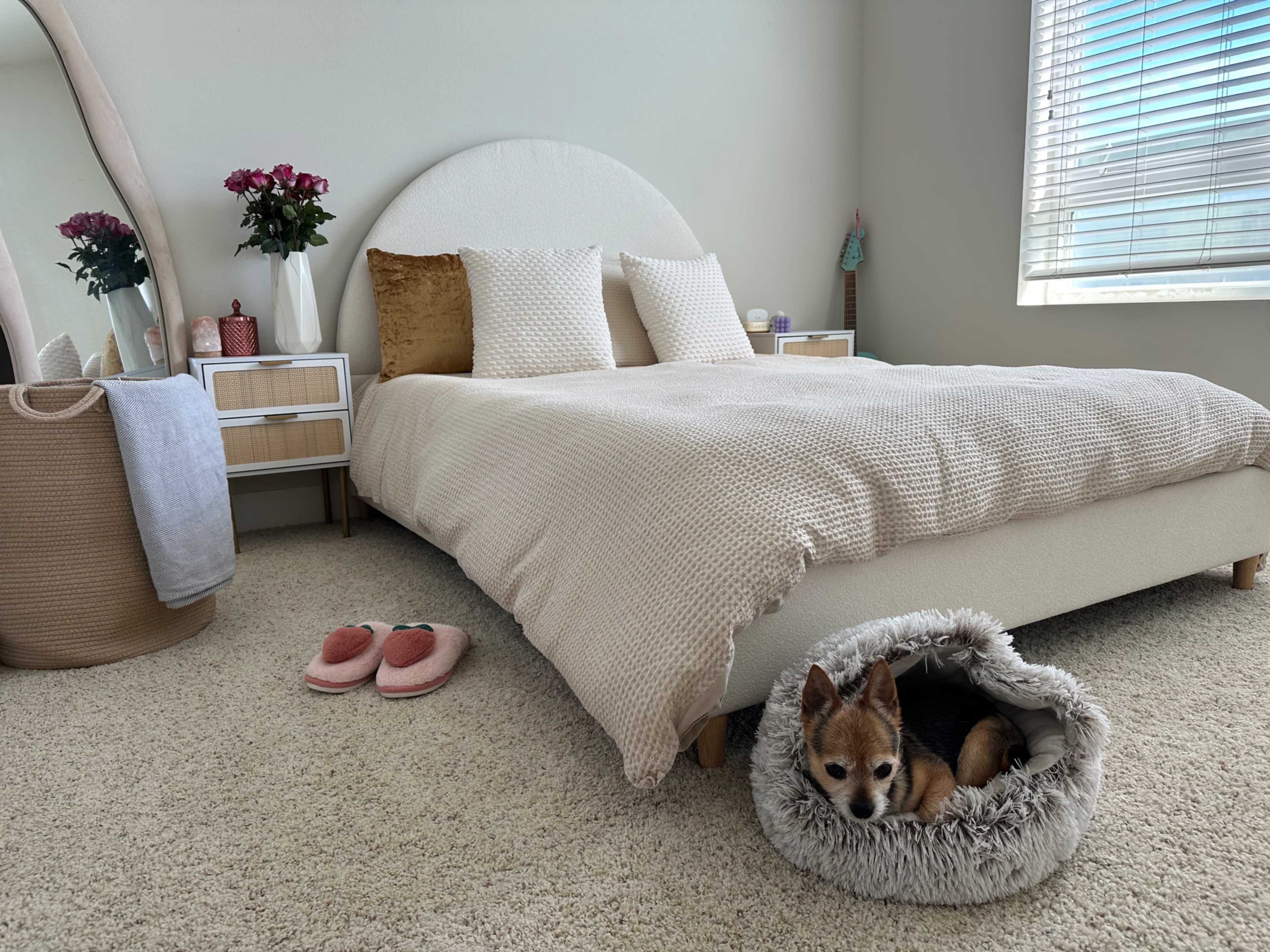 A small dog is sleeping in a cozy bed on a carpeted bedroom floor beside a light-colored bed with decorative pillows and a pair of slippers nearby.