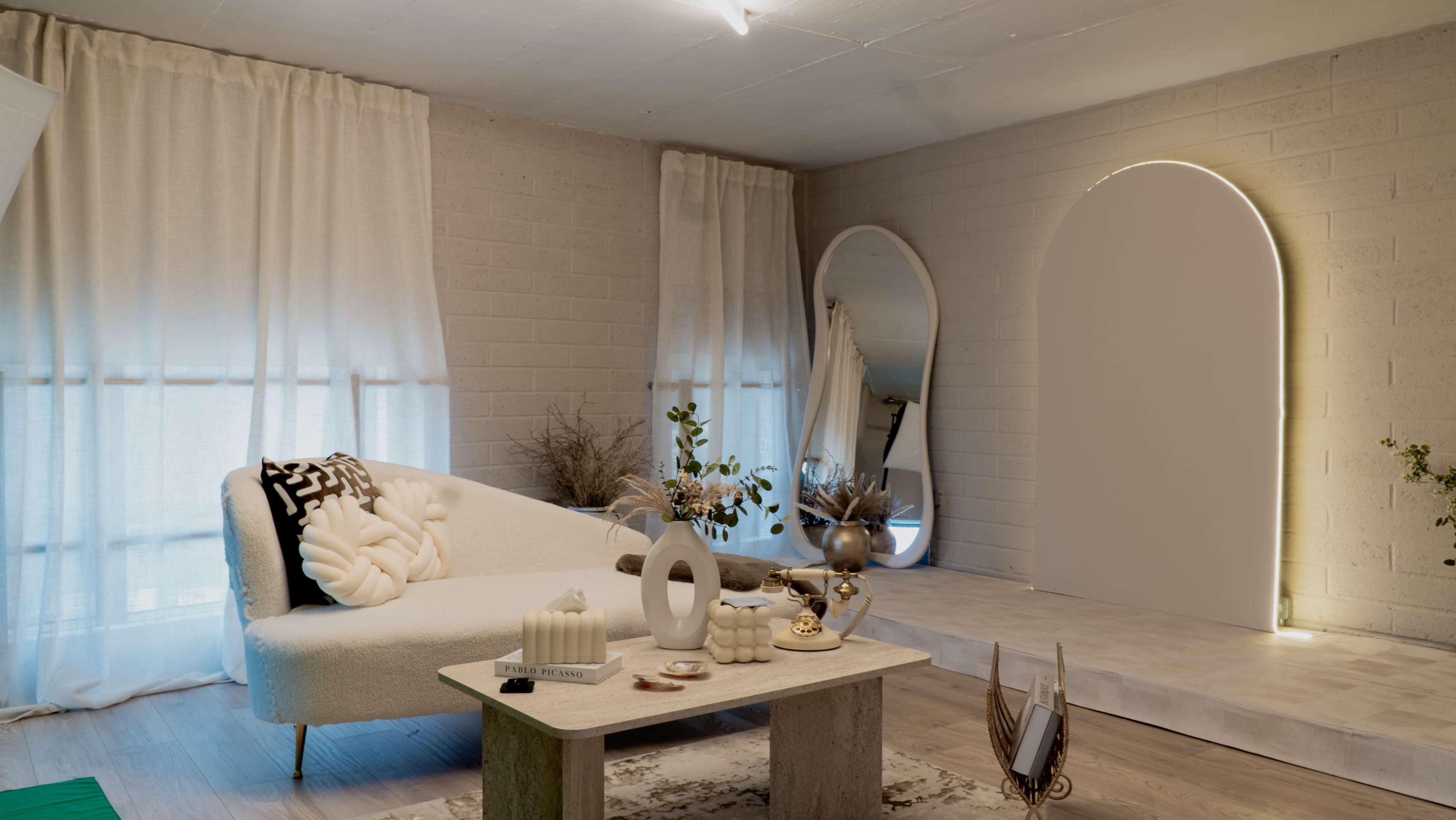 The image depicts a modern living room with a light-colored sectional sofa, a minimalistic coffee table, and decorative elements against a backdrop of soft curtains and textured walls.