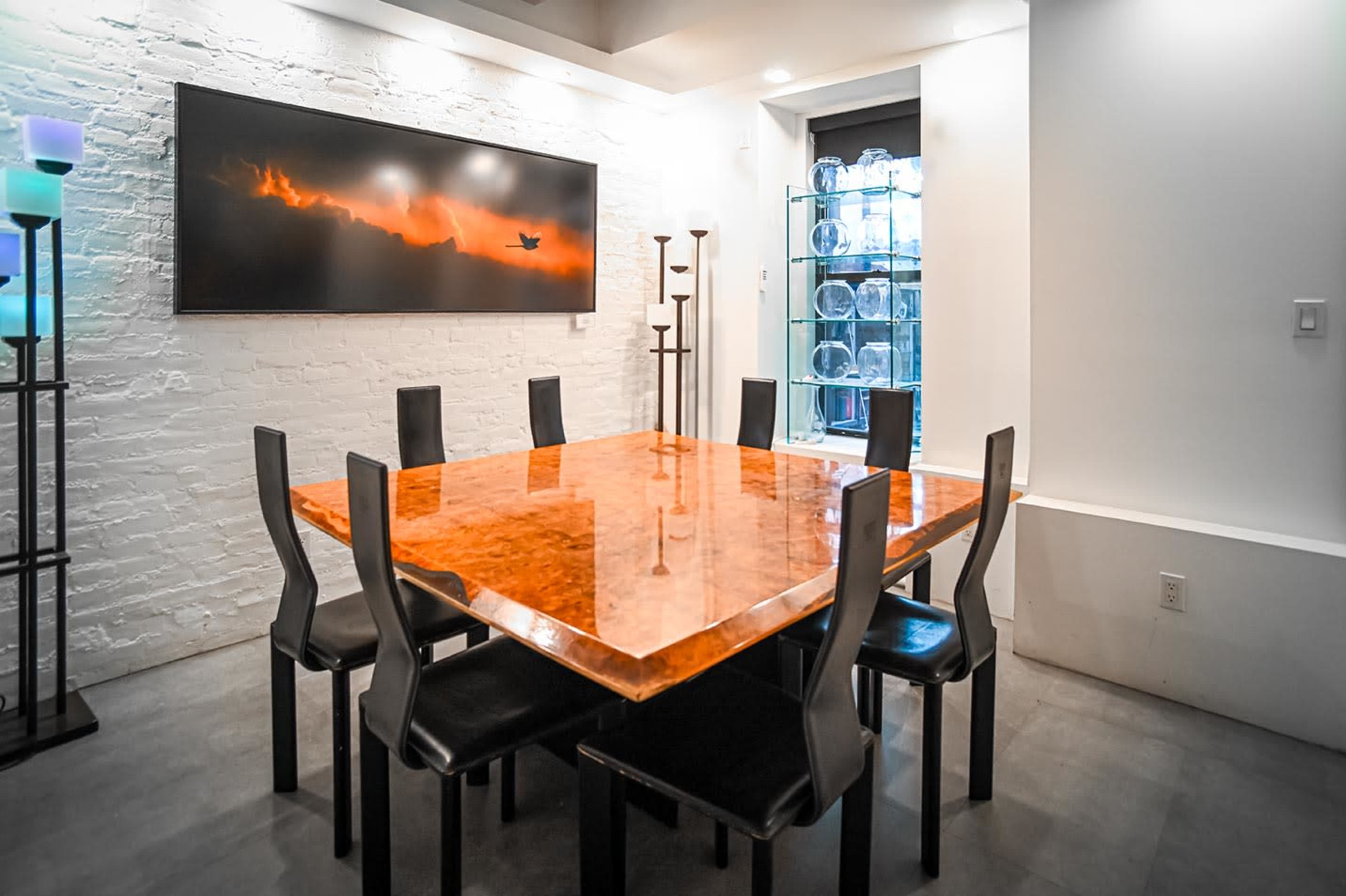 The image shows a modern dining room with a large wooden table surrounded by black chairs, next to a glass display cabinet and a mounted artwork.