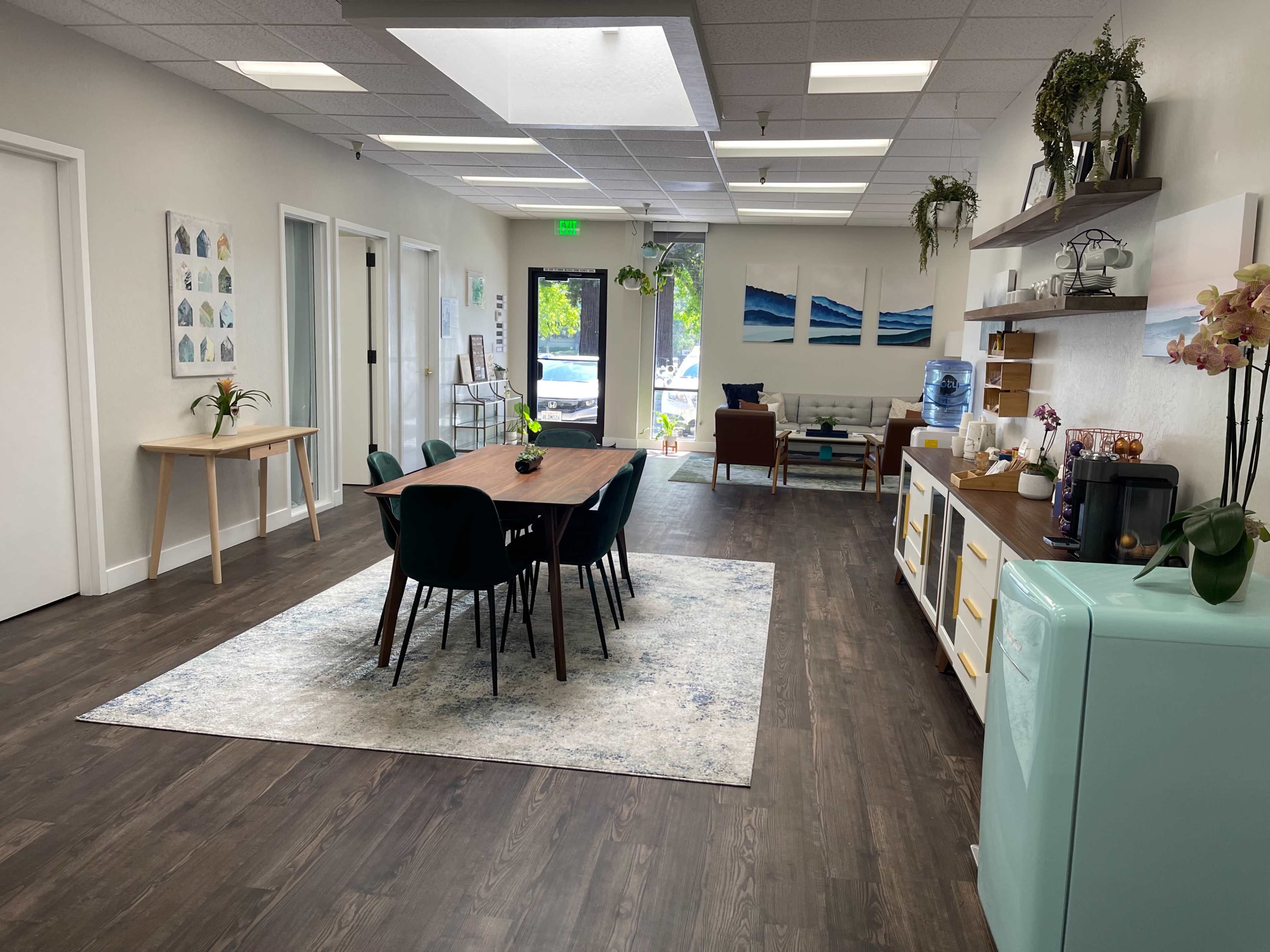 A modern office space with a dining table surrounded by chairs, a kitchenette, and a reception area featuring decorative plants and artwork.