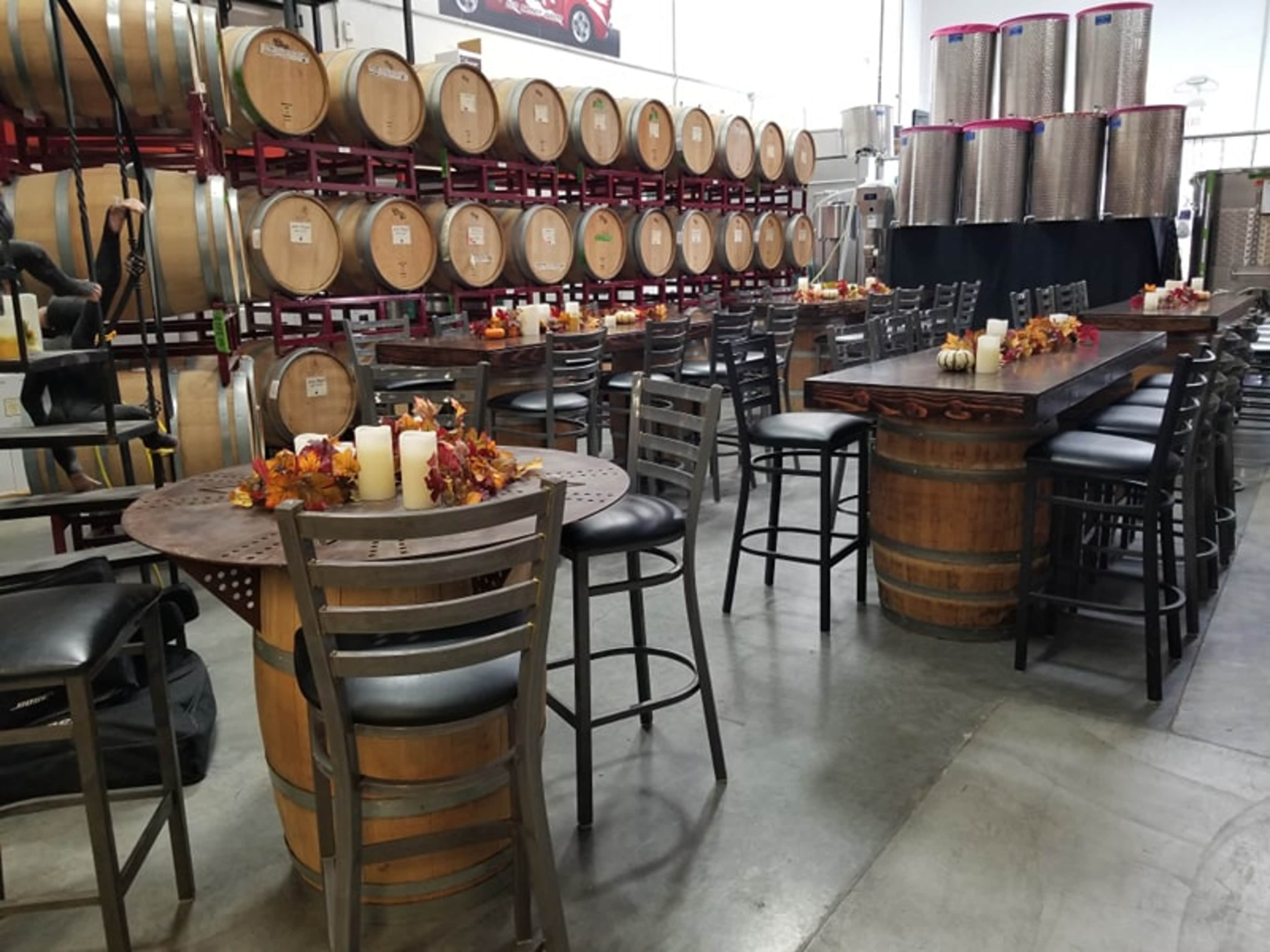 A spacious winery interior with wooden barrels lined against the wall, and tables made from barrels featuring black chairs and decorative candles.