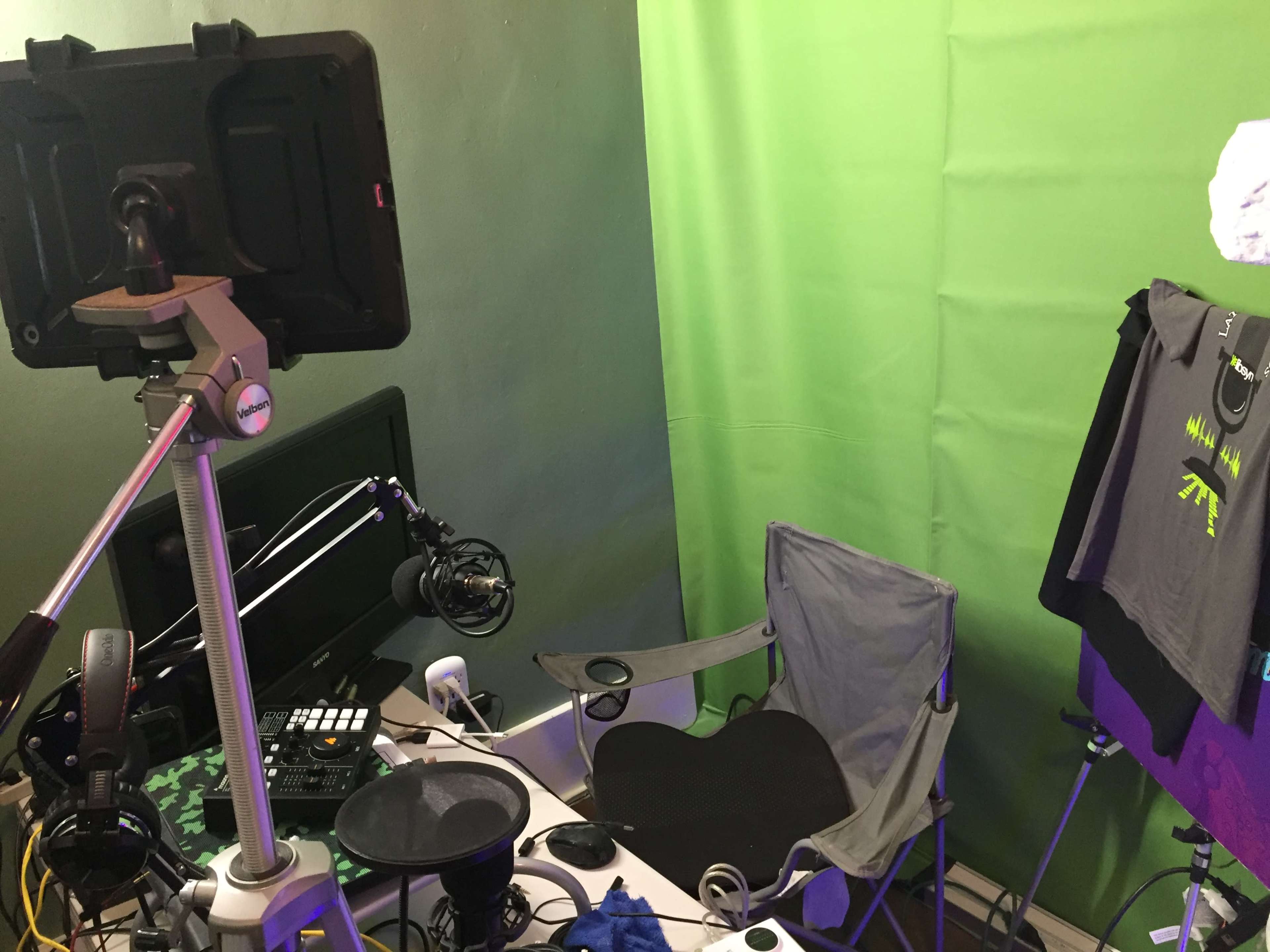 A small studio setup featuring a green screen, a microphone on a boom arm, a computer monitor, a folding chair, and clothing hanging on a rack.